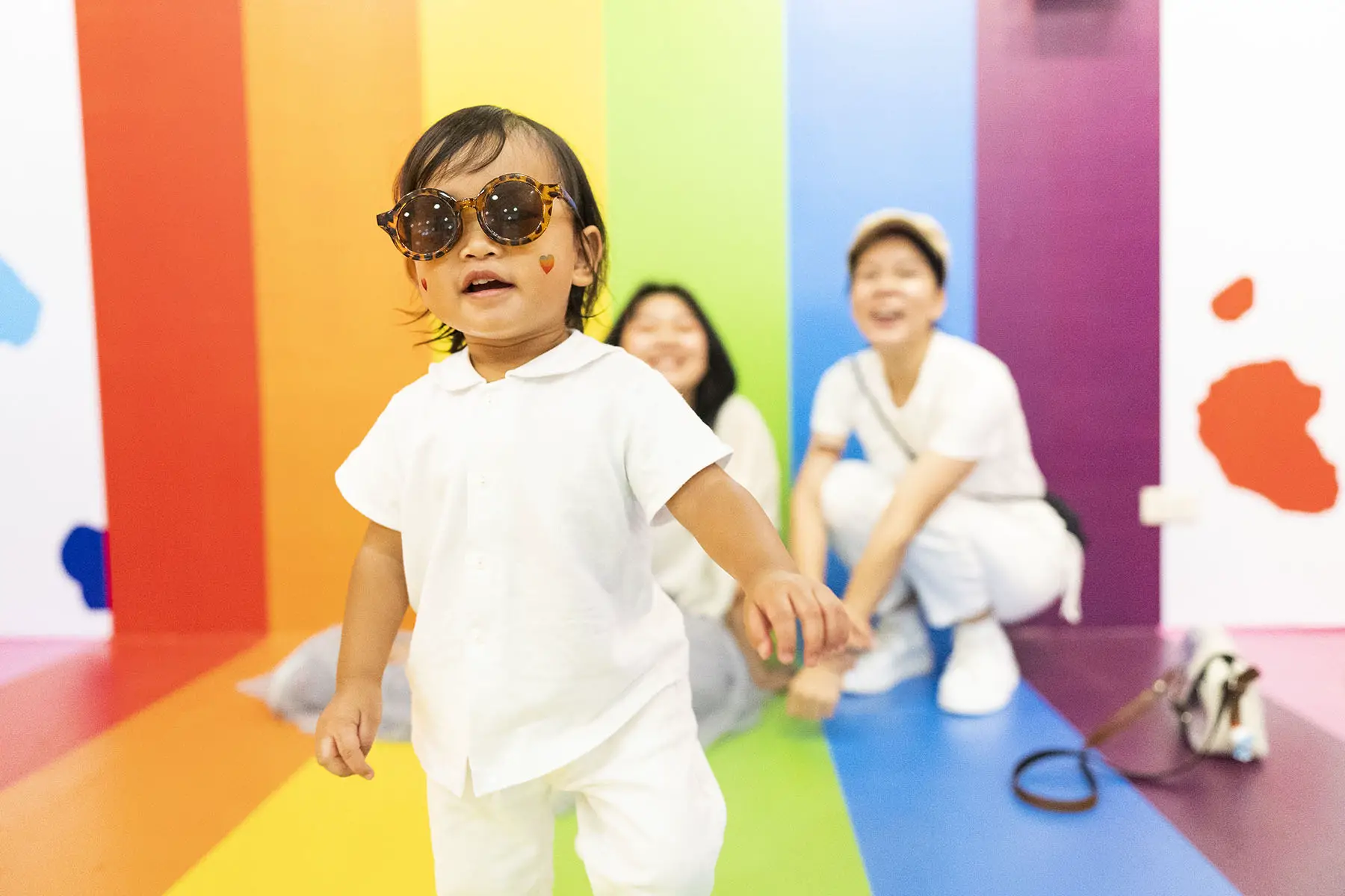 Cute baby is wearing sunglasses and walking towards the camera, their LGBT+ parents are smiling in the background in front of a rainbow flag.