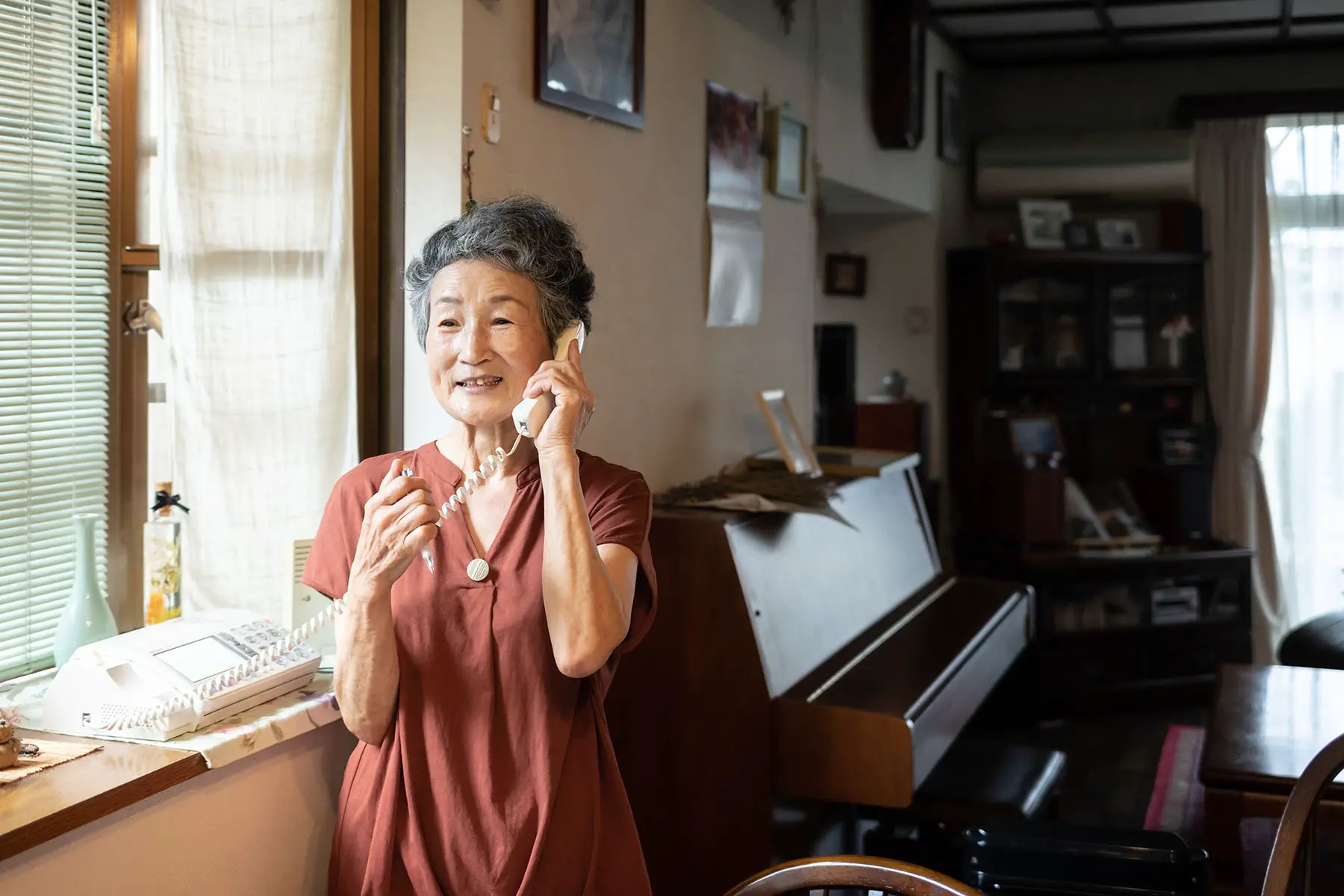 Old woman standing near a window and speaking on a landline telephone.