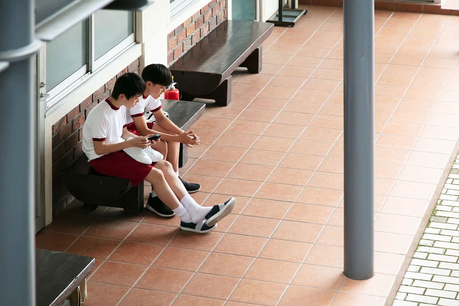 Two Japanese junior high school students sitting on a bench in the hallway, talking to each other.