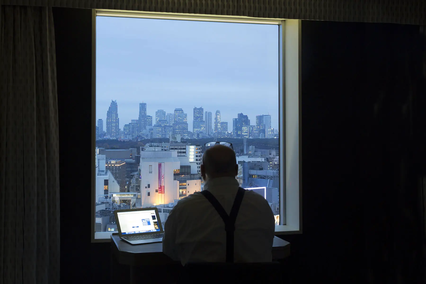 Back of a man working late while staring out of the window, looking at the Shinjuku business district in Tokyo, Japan.