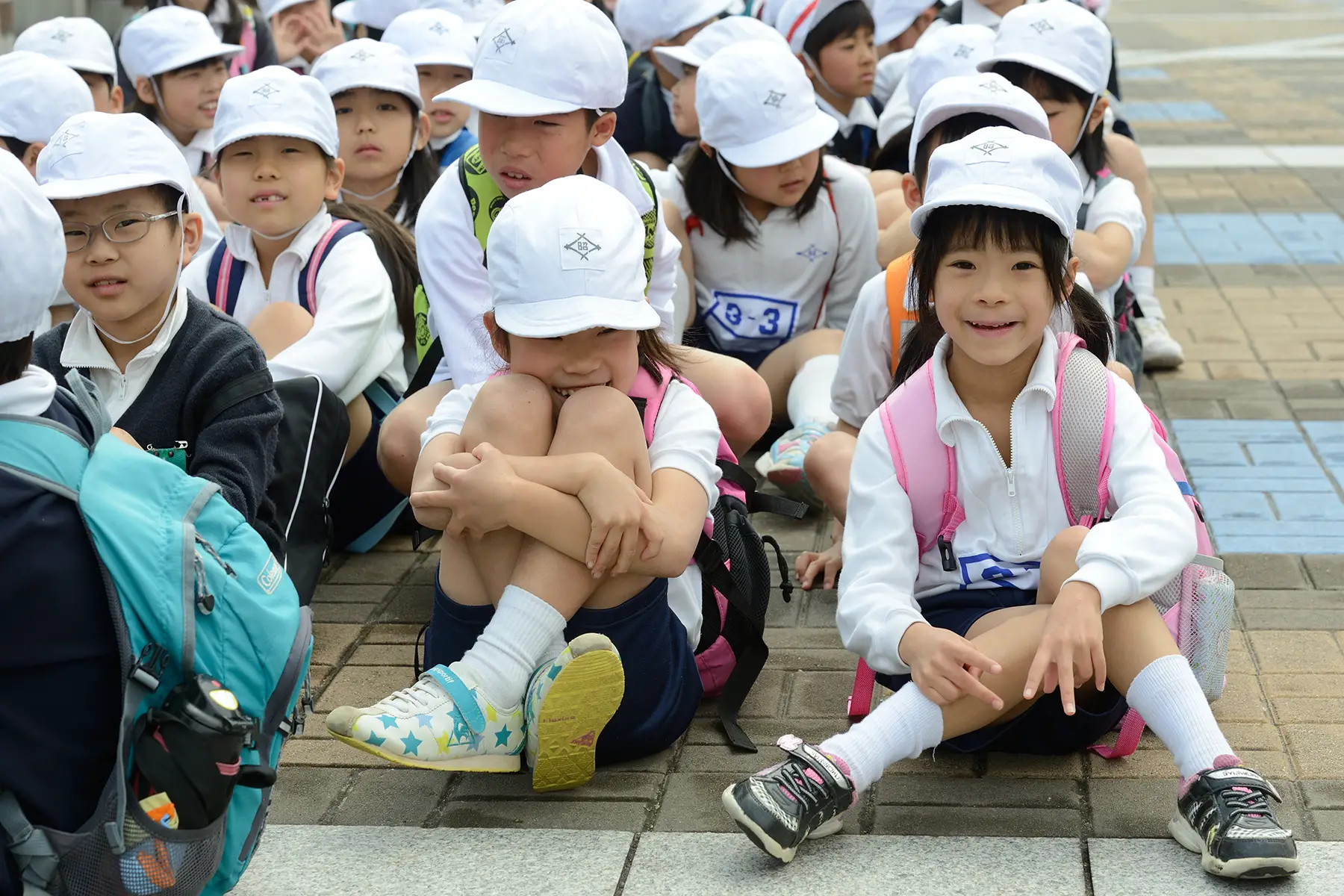 A group of primary school aged Japanese children, in uniform, sitting in a group on the ground in Osaka