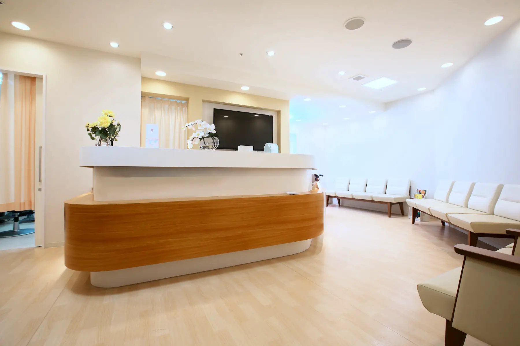 The bright, empty waiting room of a Japanese medical clinic or hospital, with flowers on the reception desk