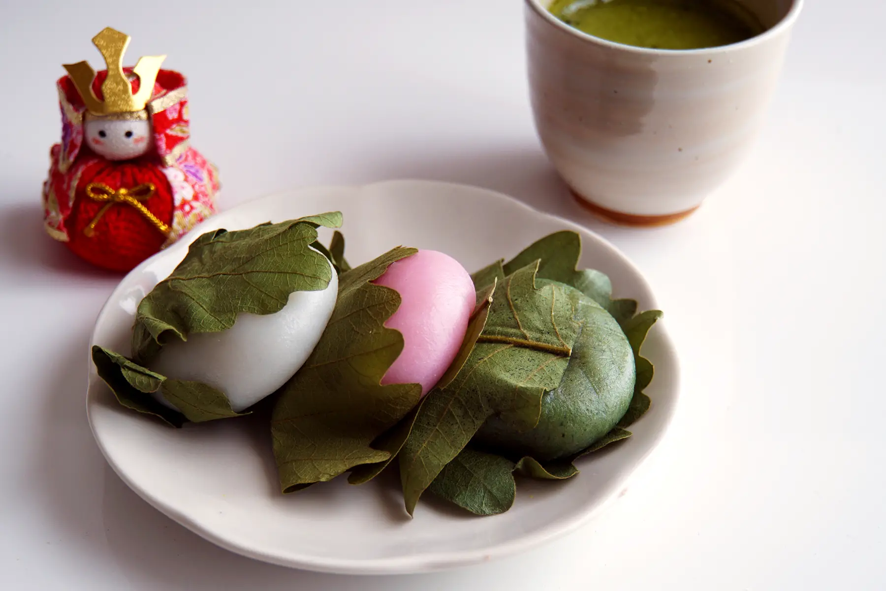 A plate of three rice cakes in white, pink, and green, wrapped in leaves. Accompanied by a statuette and cup of tea.