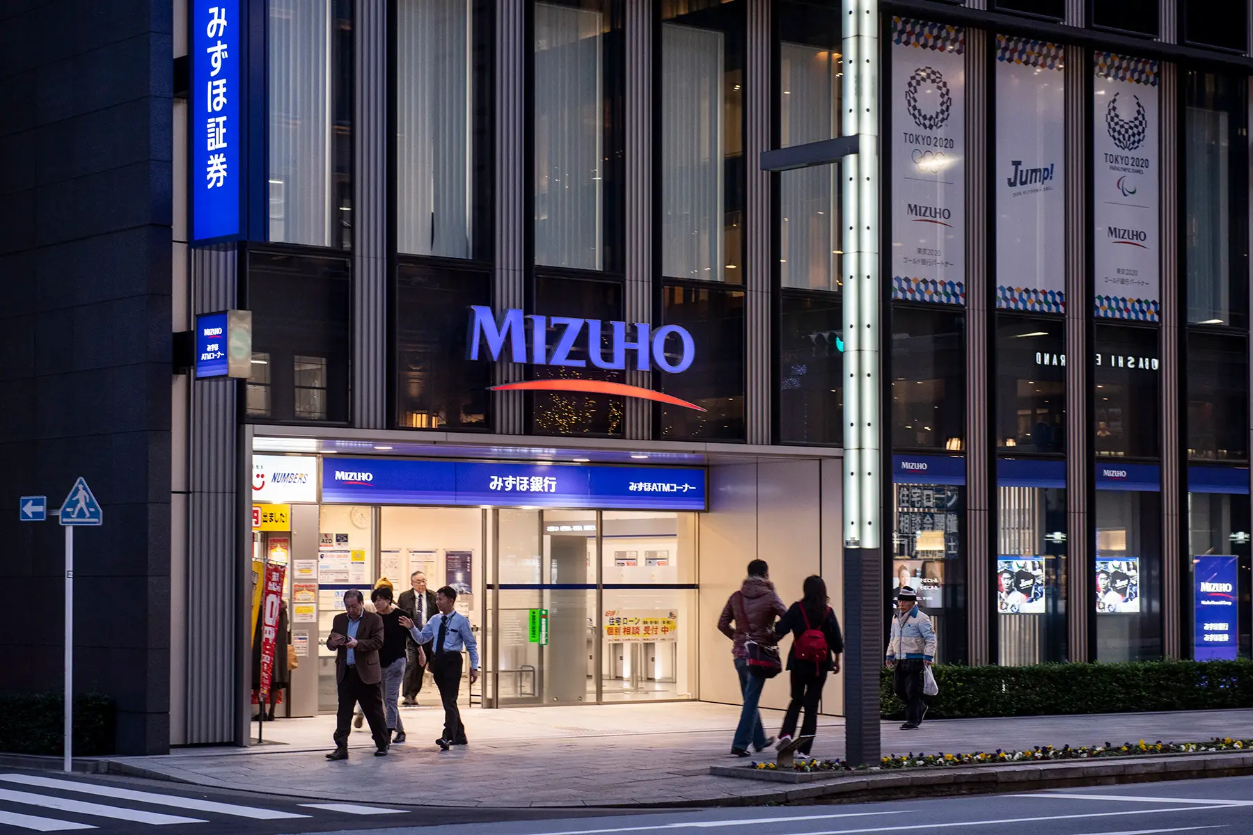 Mizuho bank at night with lights on and people walking past
