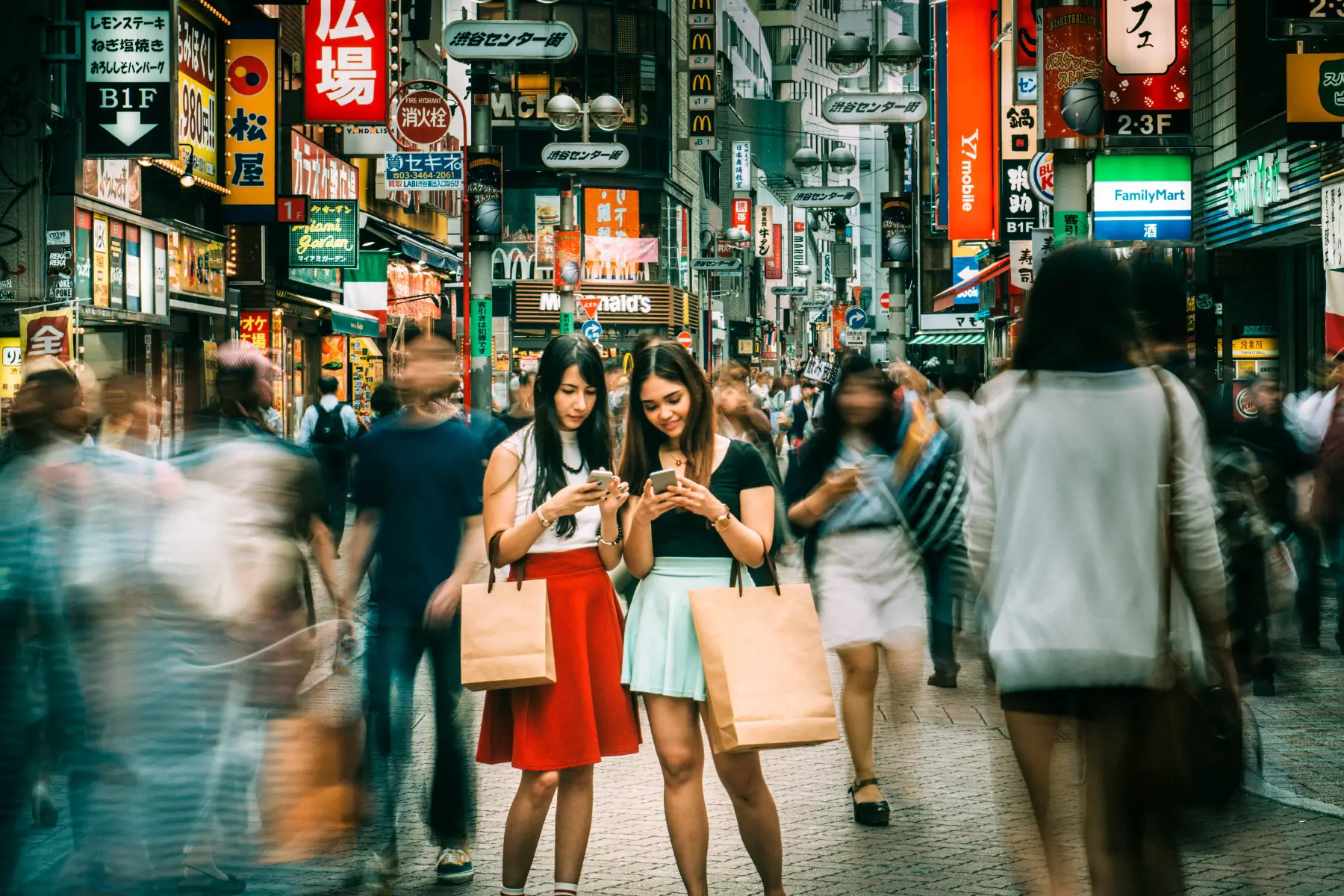 two Japanese teenage girls standing in the streets of Shibuya in Tokyo holding shopping bags and looking down at their smartphone