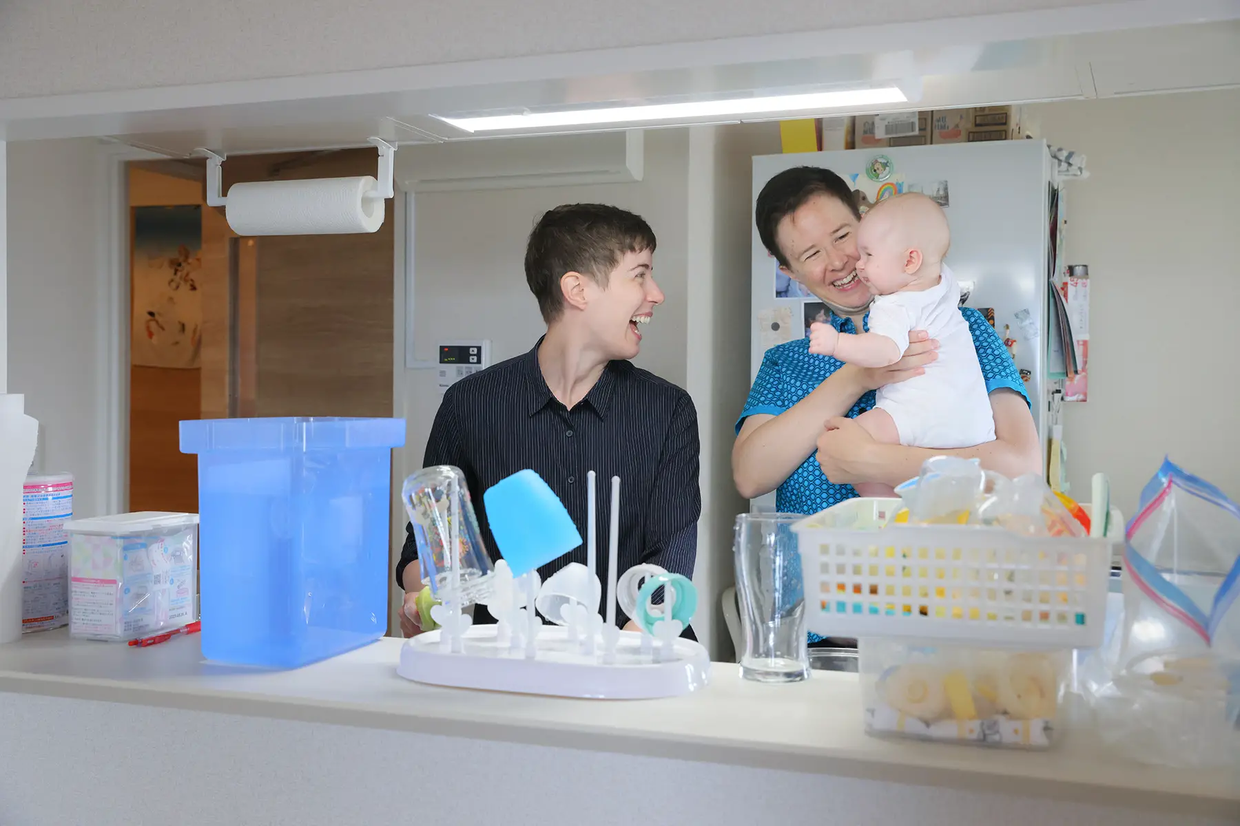 Two mums (a couple) standing in a kitchen preparing a bottle, one hold their baby, both looking and laughing at their baby 