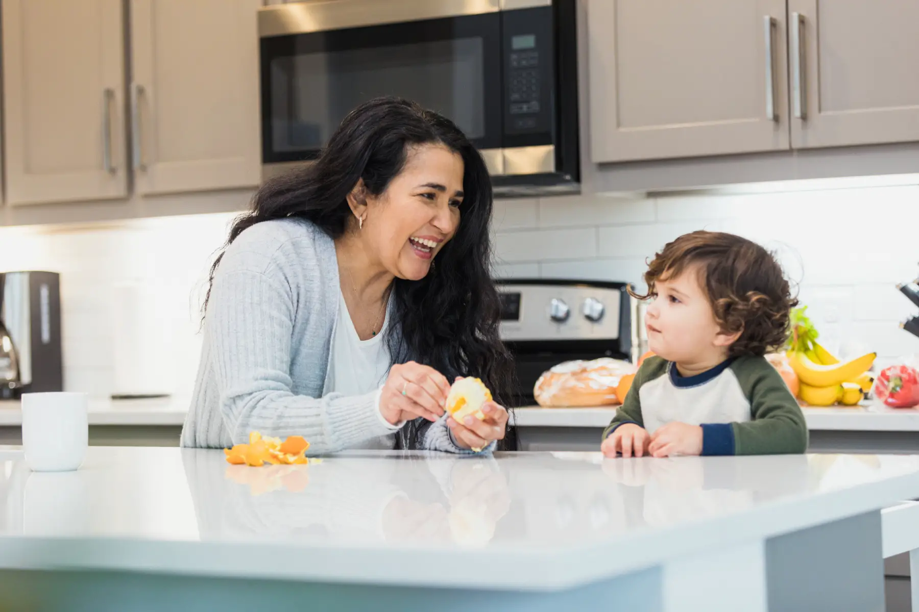 a nanny smiling and feeding fruit to a happy toddler in the family kitchen