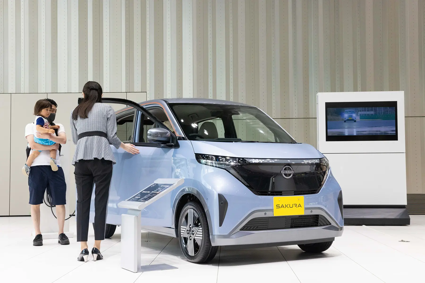 A salesperson shows a new Nissan Sakura to a child and its parent. The car is boxy and light blue.