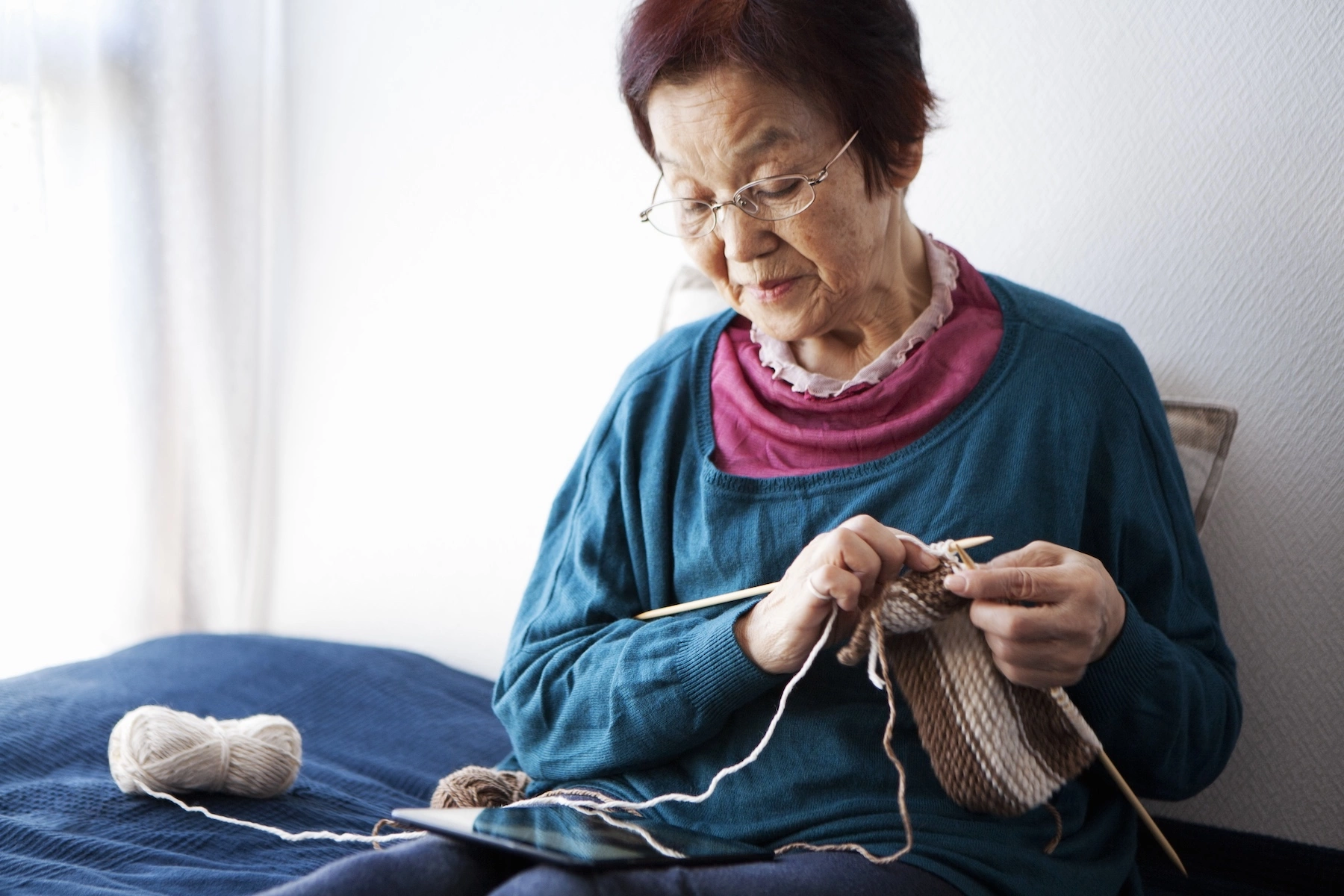 An older adult woman senior woman sits in bed knitting while referring to a pattern on her tablet