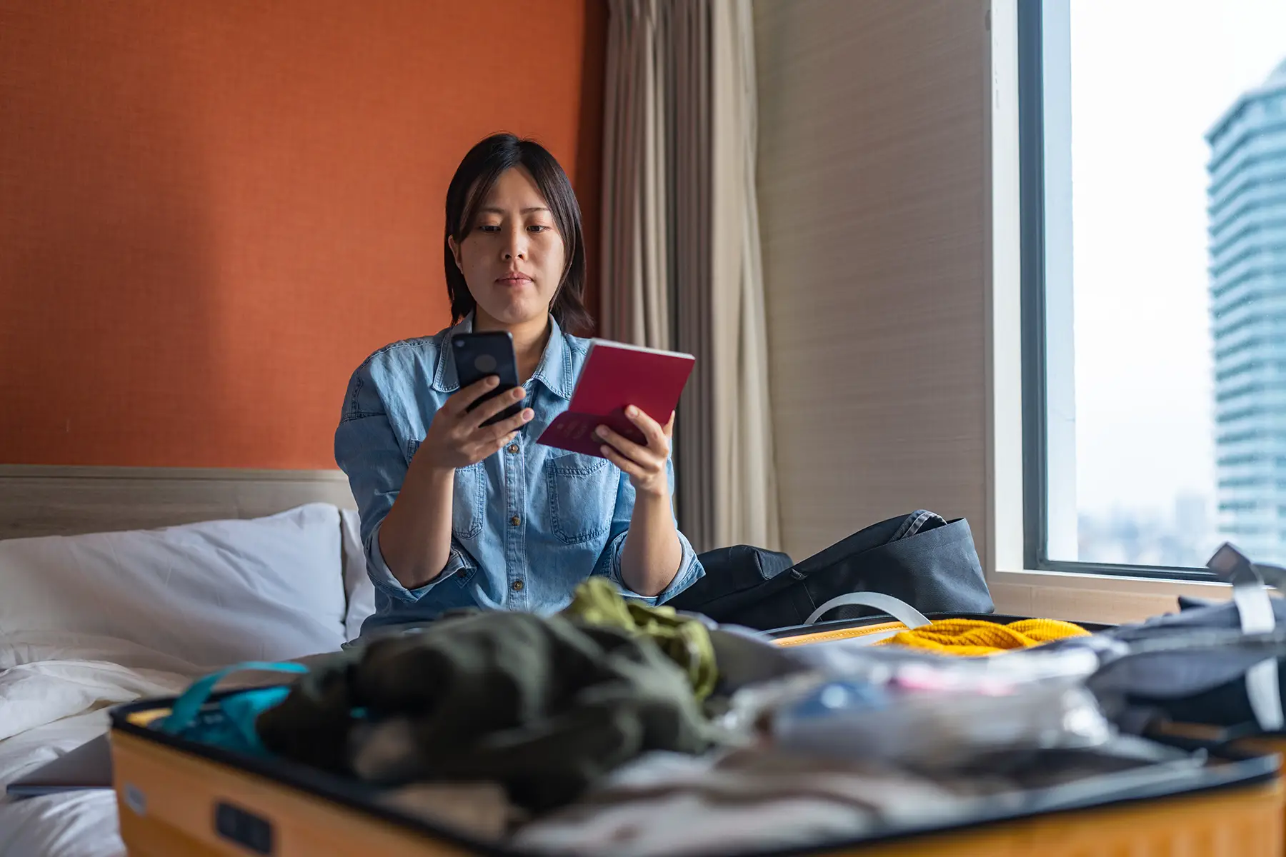 A woman sits in her bed with a  suitcase in front of her. She hold her passport open while checking something on her smart phone.