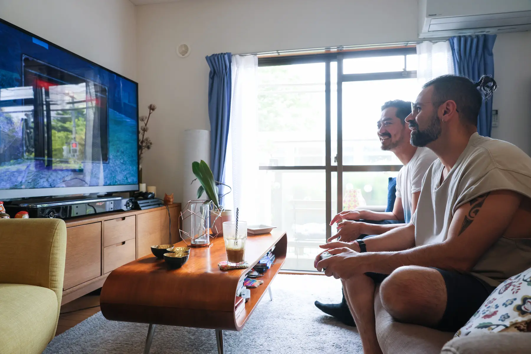 Two men sit on a couch side-by-side in front of a television playing a video game and laughing