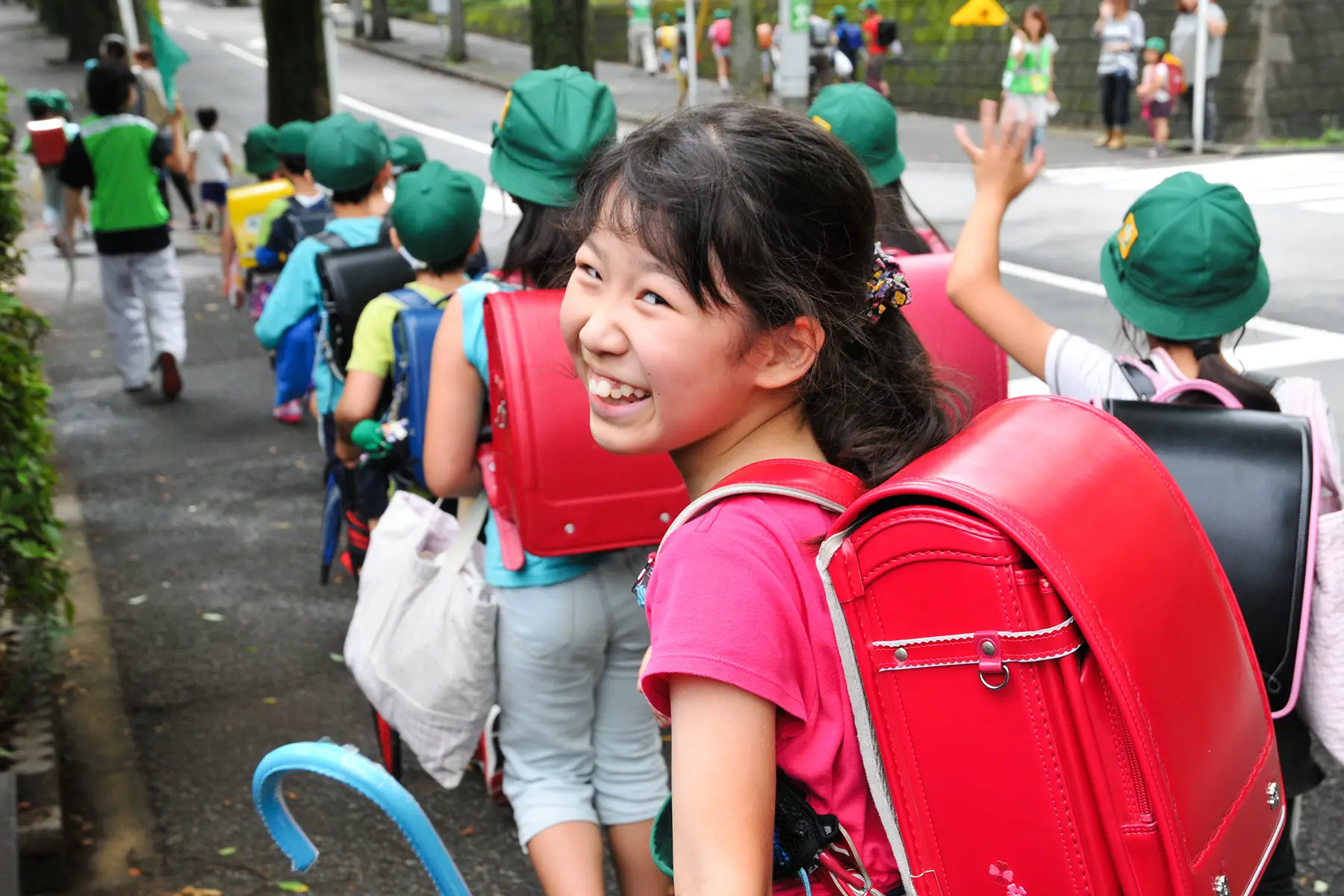 Primary school children with green hats and bright colored backpacks walk away from camera, one little girl looks around and smiles
