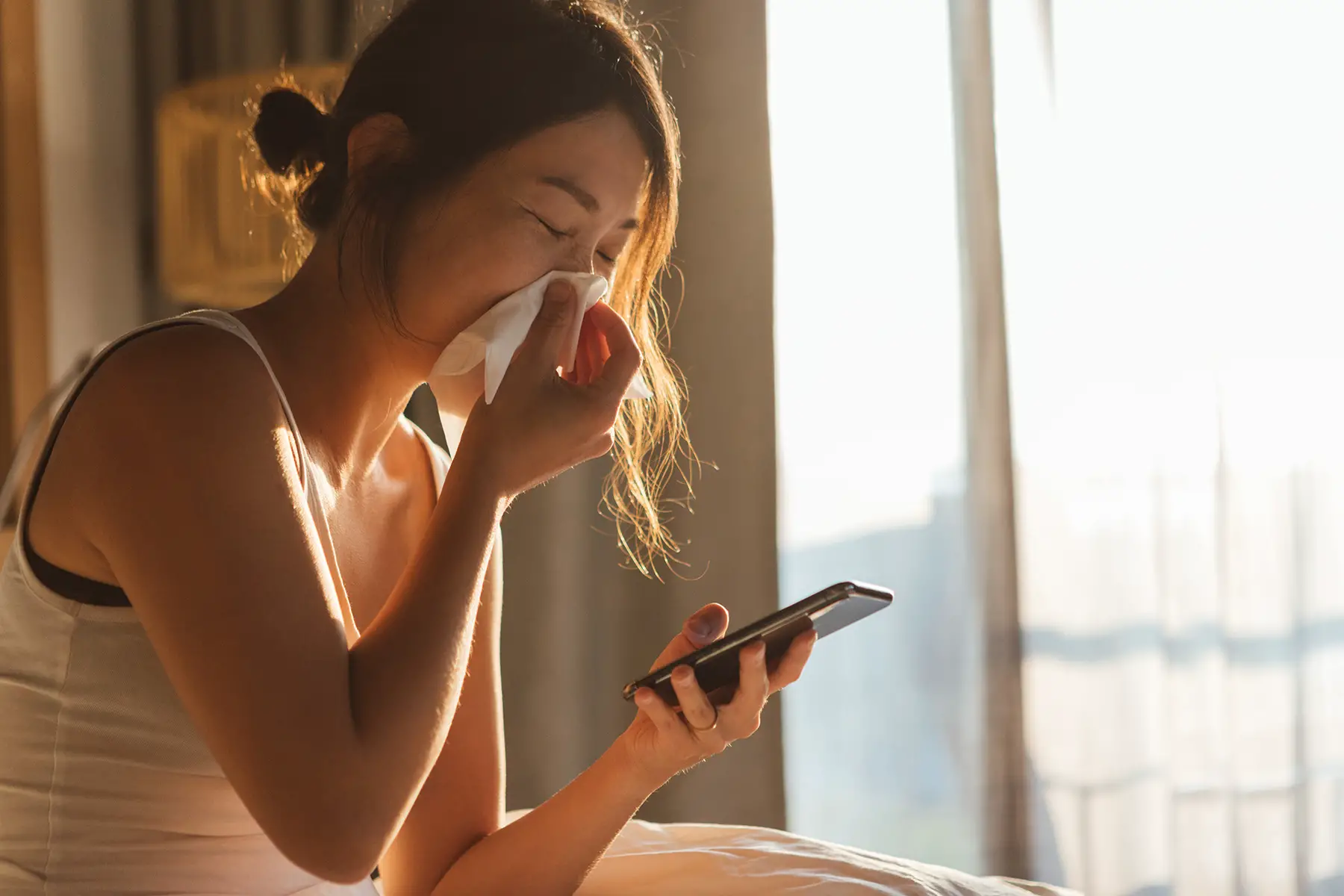 Woman sits in bed sick, blowing her nose while holding her phone