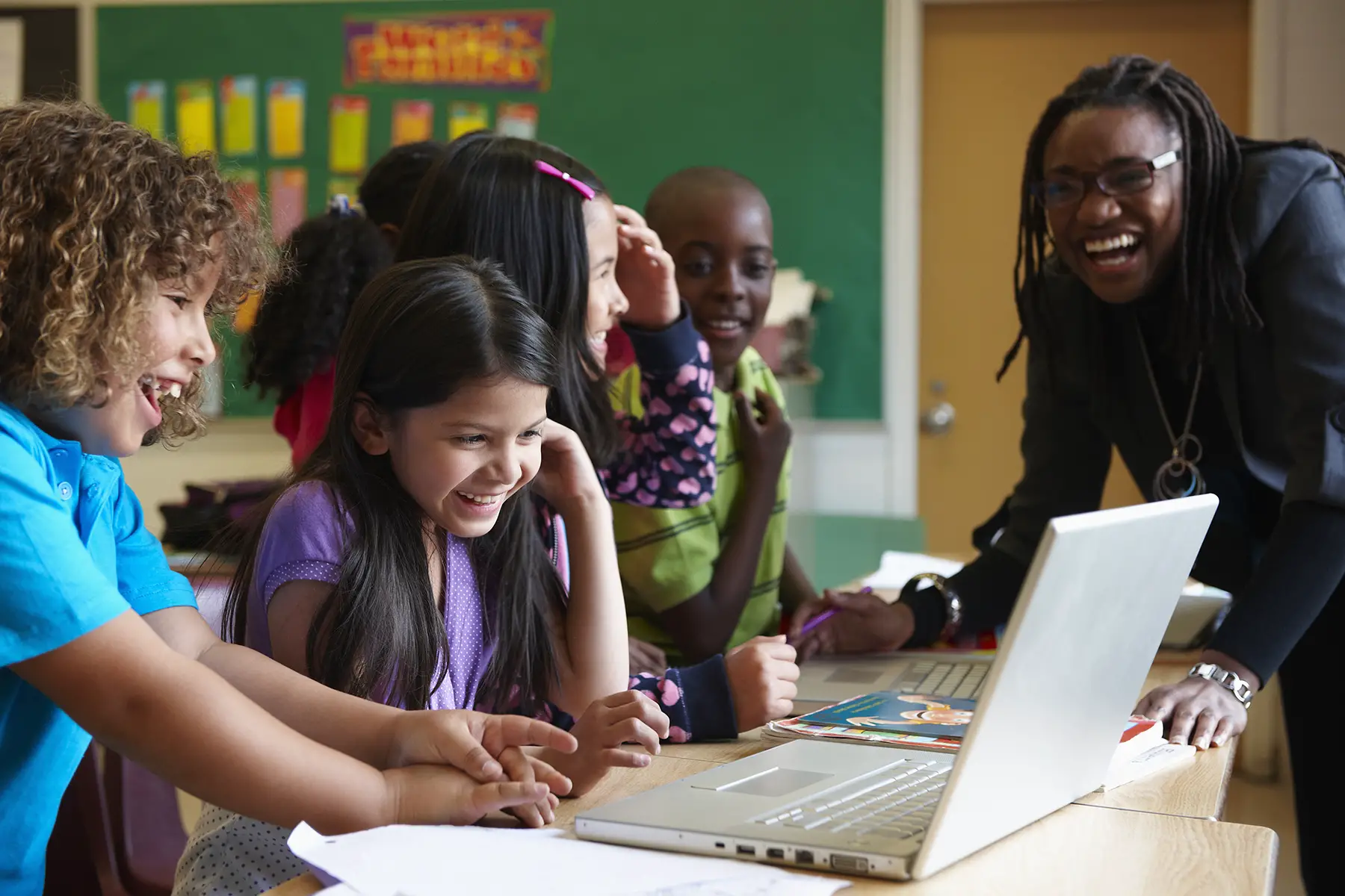 A teacher laughs with her primary school students while they are working on their laptops