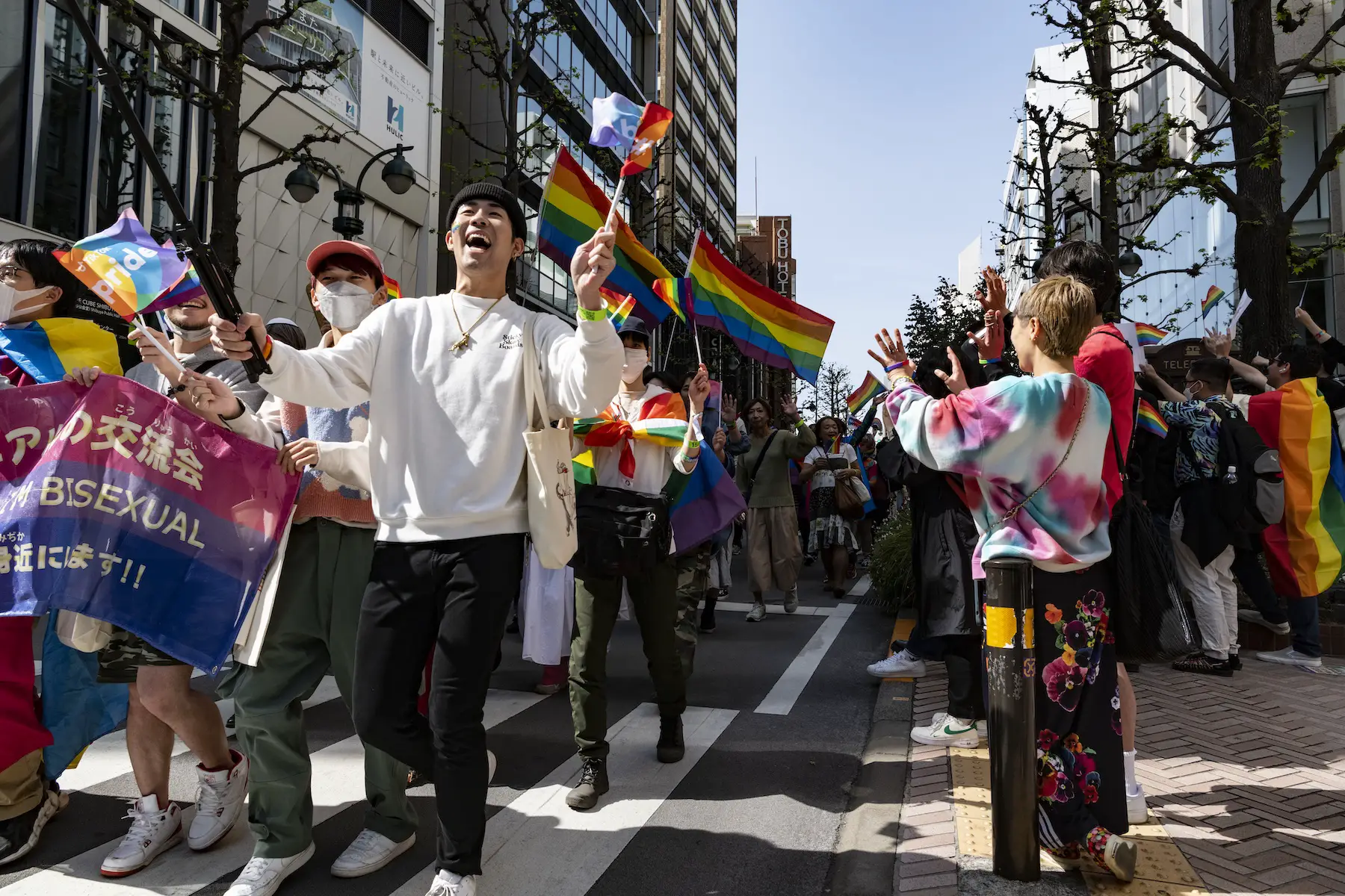 A crowd marches down the street in Tokyo, Japan during Pride 2023; everyone is waving rainbow flags and some wearing medical face masks