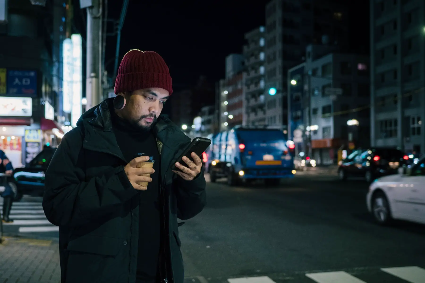 a man standing in a street in Tokyo at night, looking down at his smartphone as he requests an uber through his app