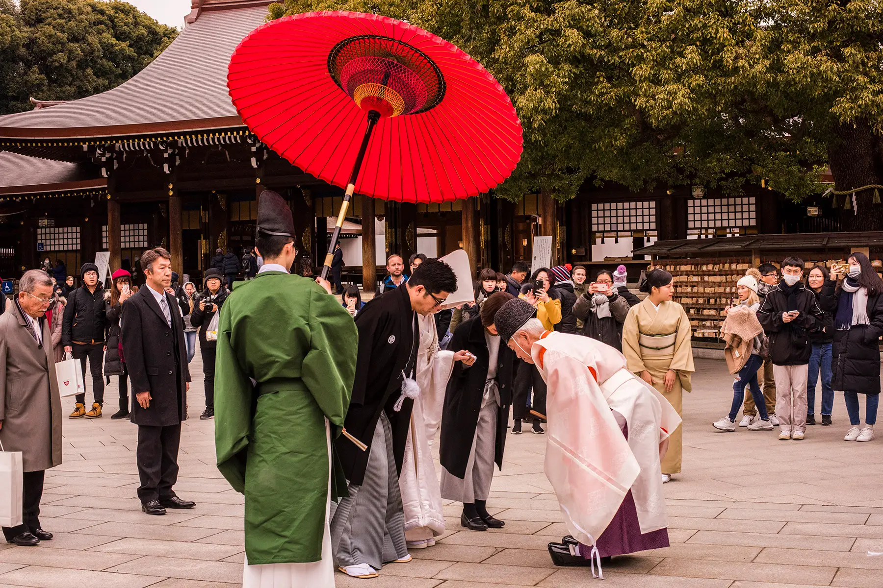A traditional Japanese wedding ceremony outside a temple. The couple stands under a red umbrella.
