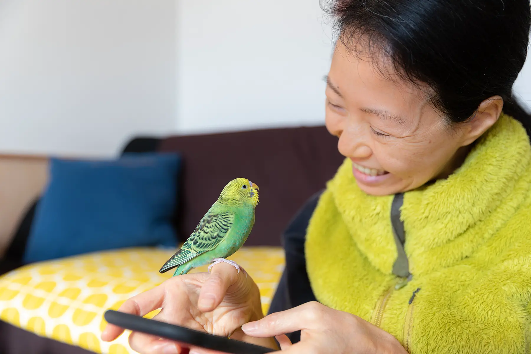 A woman using a phone, smiling at her pet parakeet which is sitting on her hand