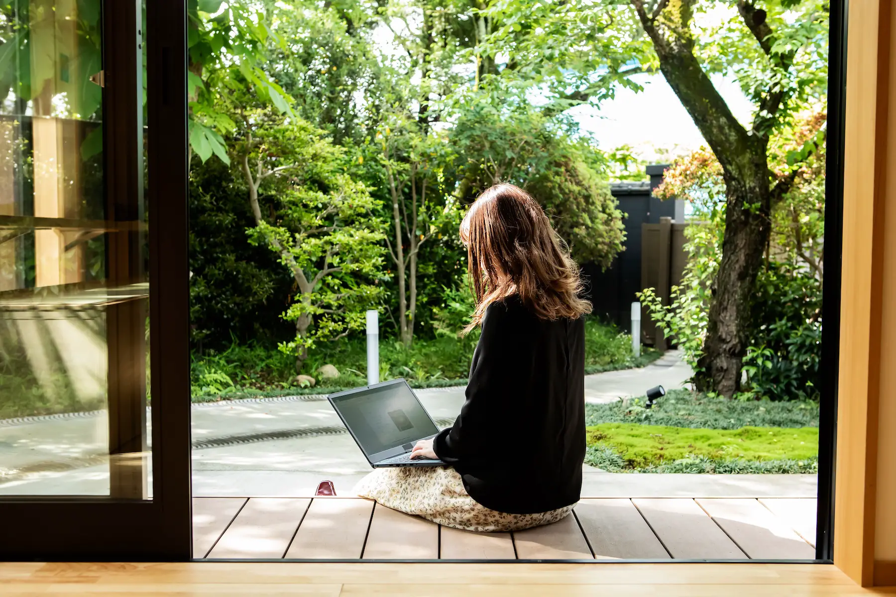 A woman sits on her porch working on a laptop. The sliding doors to her house are open and it's a sunny day.