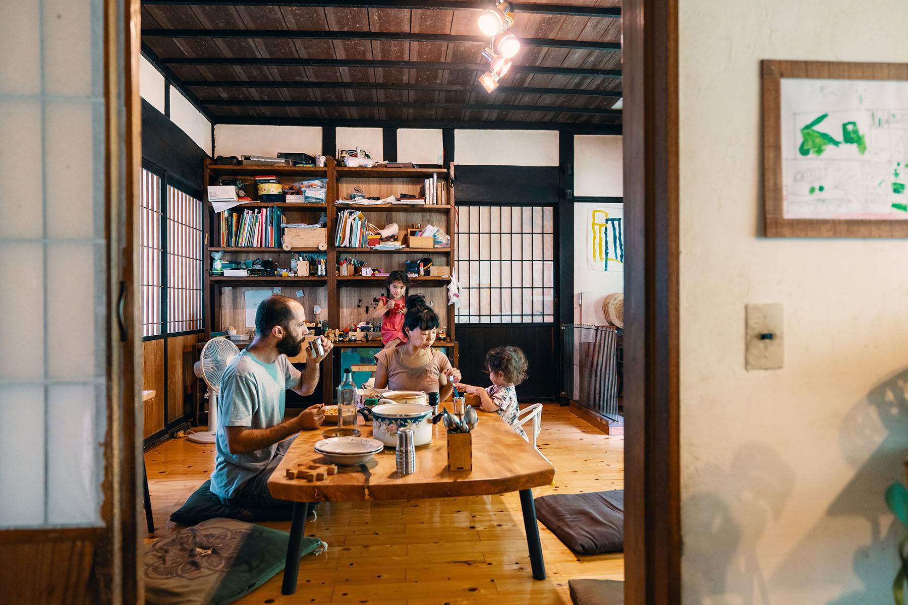 Family eating lunch in a traditional Japanese house in rural Japan. Father, mother, and little baby are sitting on the floor, at the table. Young daughter is standing closeby near the book shelves.