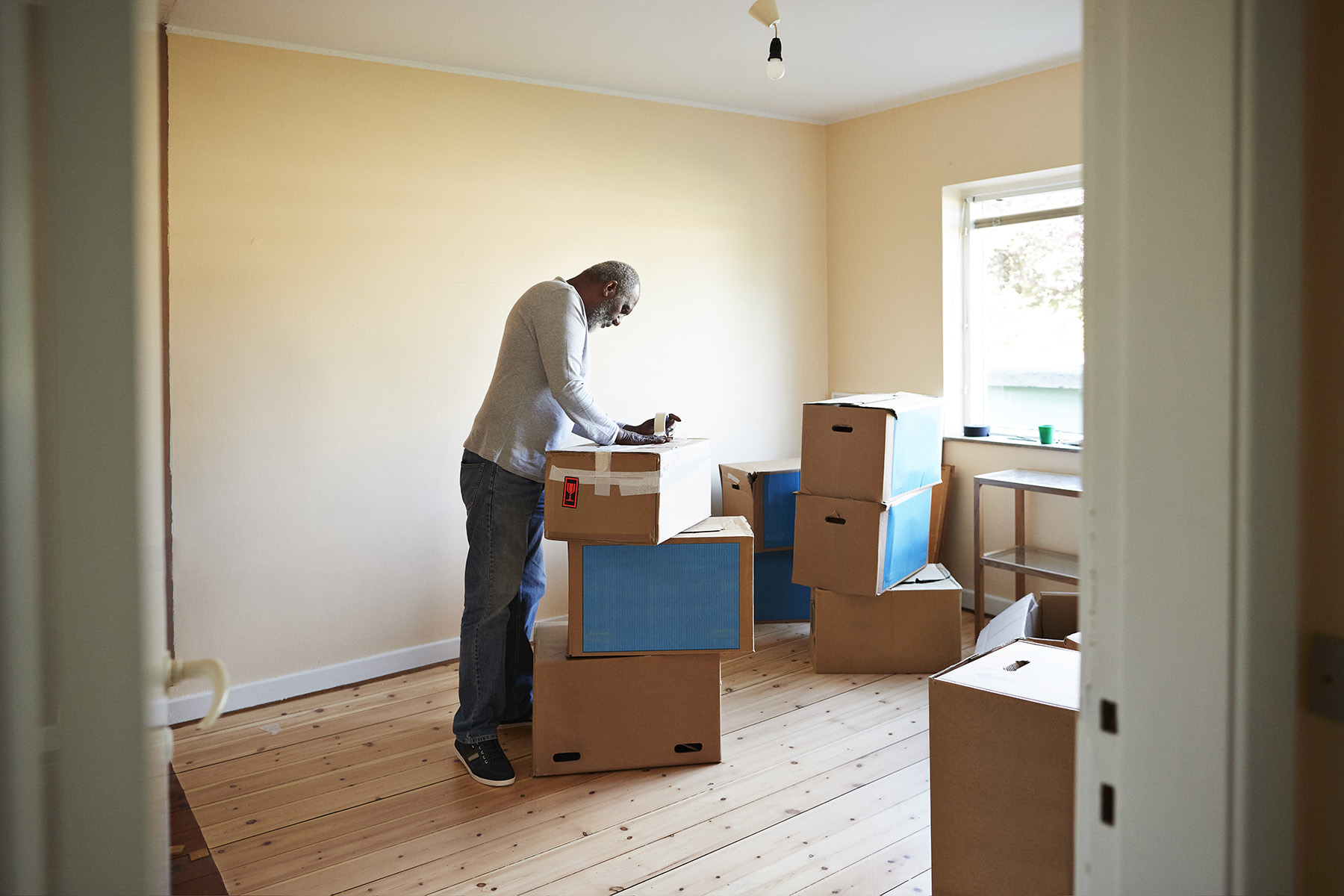 Man standing in an empty room (except for moving boxes), taping a box while getting ready to relocate. 