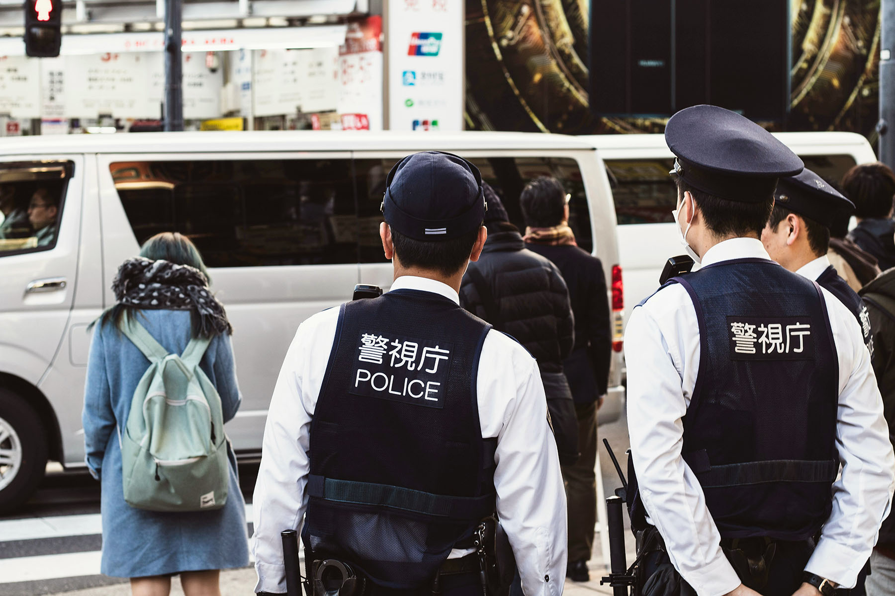 Two police officers standing on the street, with their backs turned towards the camera. They're looking at a woman with a backpack.