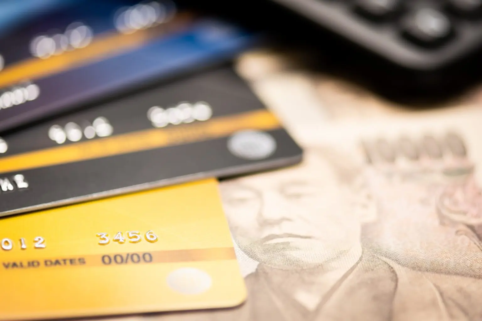 A close-up, shallow-depth-of-field shot of several credit cards fanned out on top of a Japanese 10,000 yen banknote. The foreground features a gold credit card with embossed numbers, while the background shows a blurred calculator and other dark-colored cards.