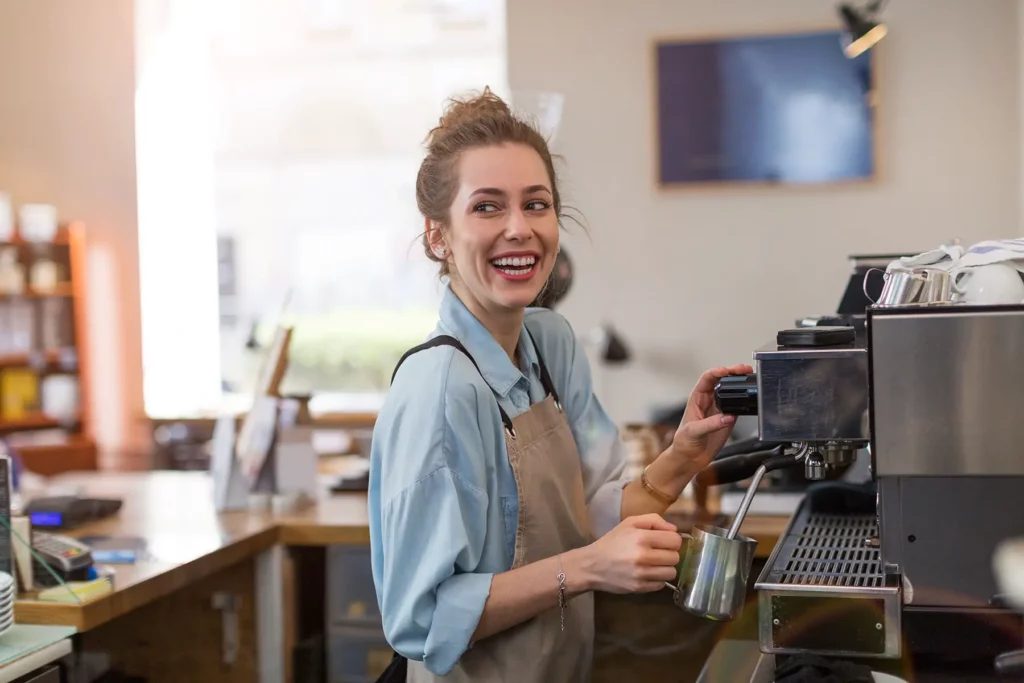 Young barista preparing coffee for customers at her cafe counter, she has a huge smile.