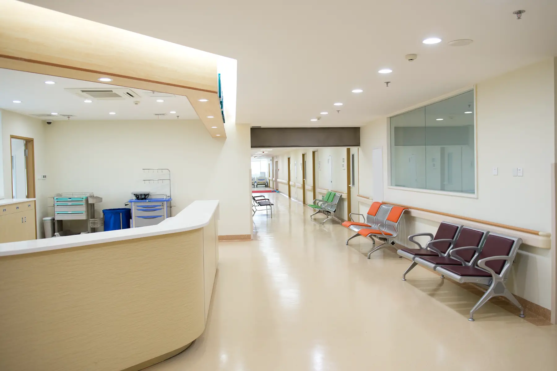 An empty hospital corridor, with chairs on one side and a nurses' station on the other