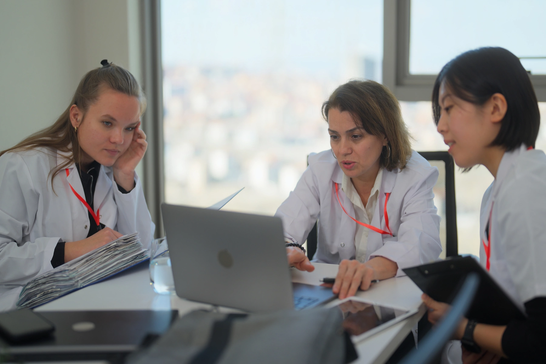 A group of three female doctors sit around a table looking at a laptop during a staff meeting