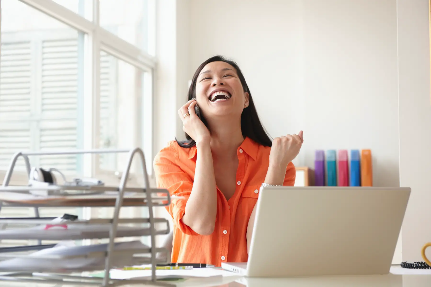 an Asian woman chatting on the phone in her office and looking happy and excited