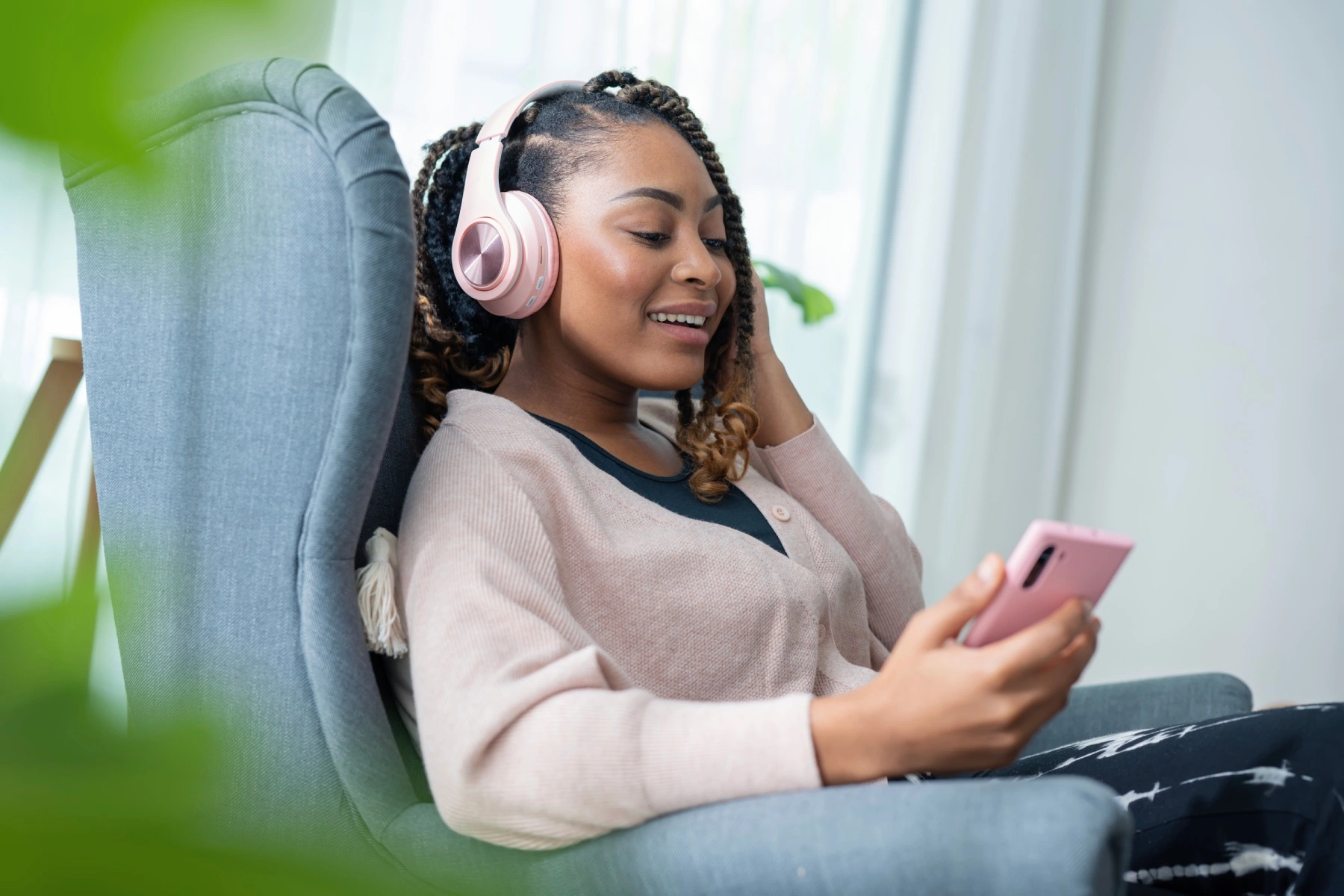 a woman of color relaxing in an armchair and wearing headphones as she looks down at her smartphone in her hand