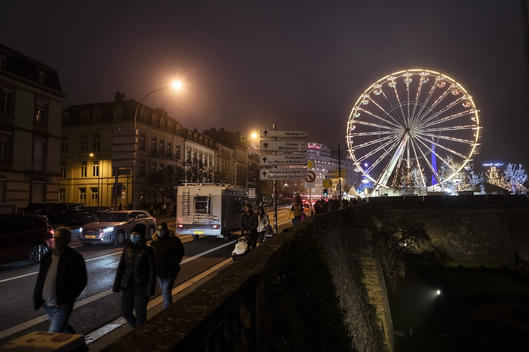 Pedestrians walk along the city walls of Luxembourg at night with the Christmas market lights and ferris wheel in the background