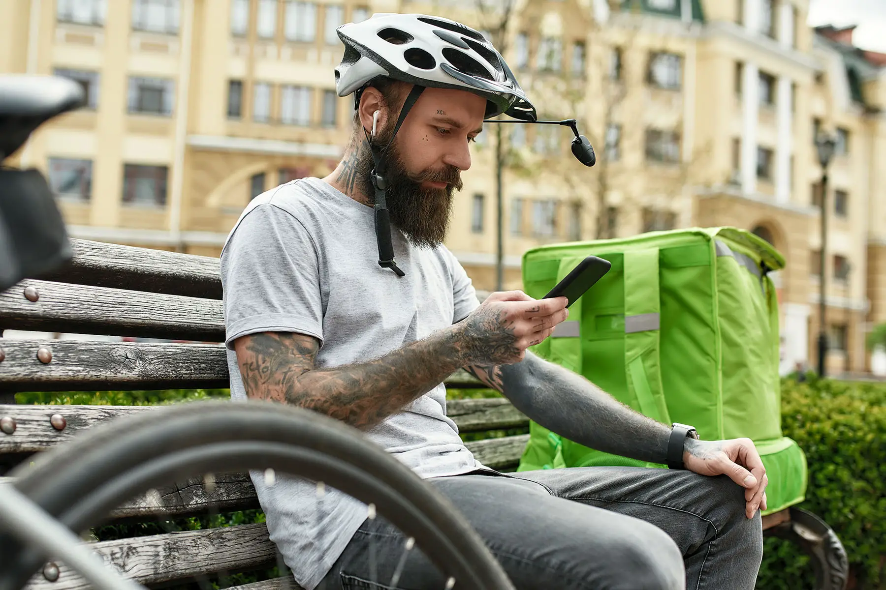 Tattooed delivery man with a helmet checking his phone while sitting on the bench outdoors. He has a green  thermo bag  next to him.