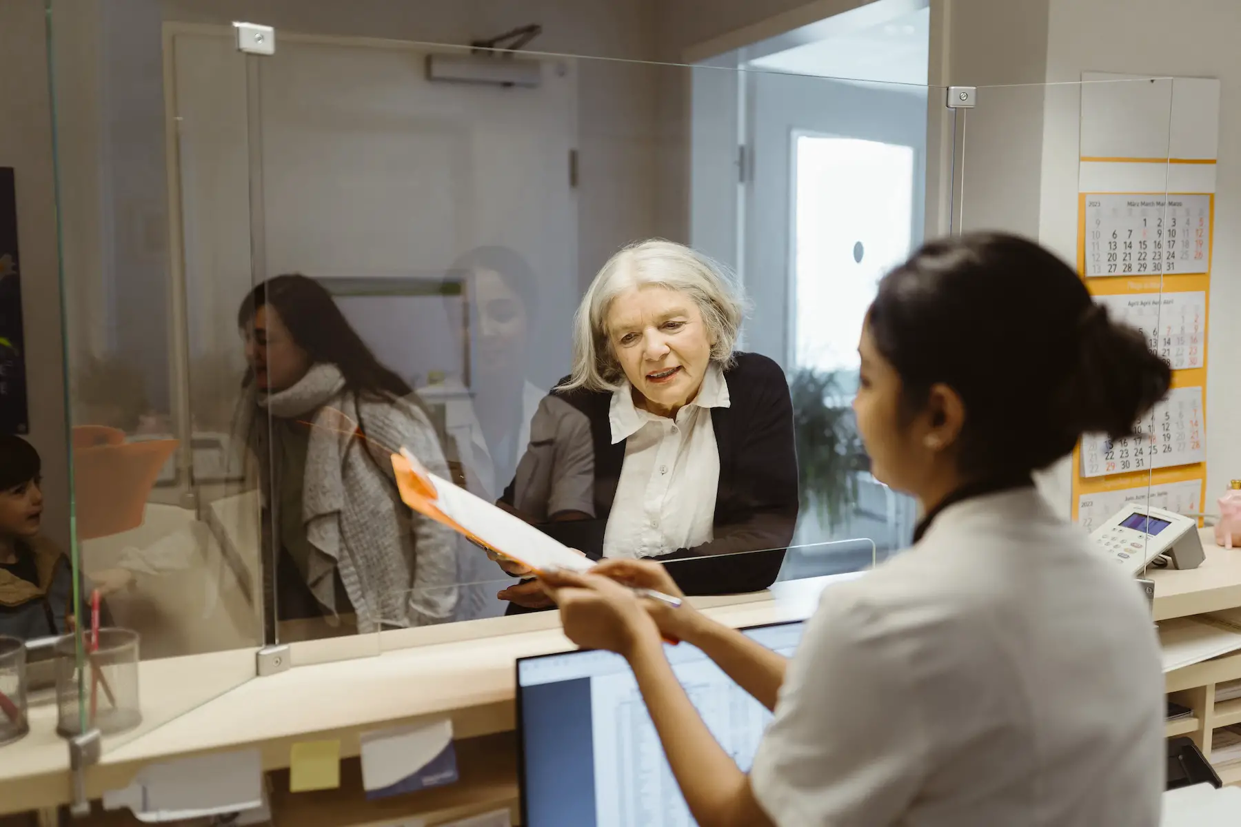 An older patient prepares to pay her medical bill with the receptionist who stands behind a desk