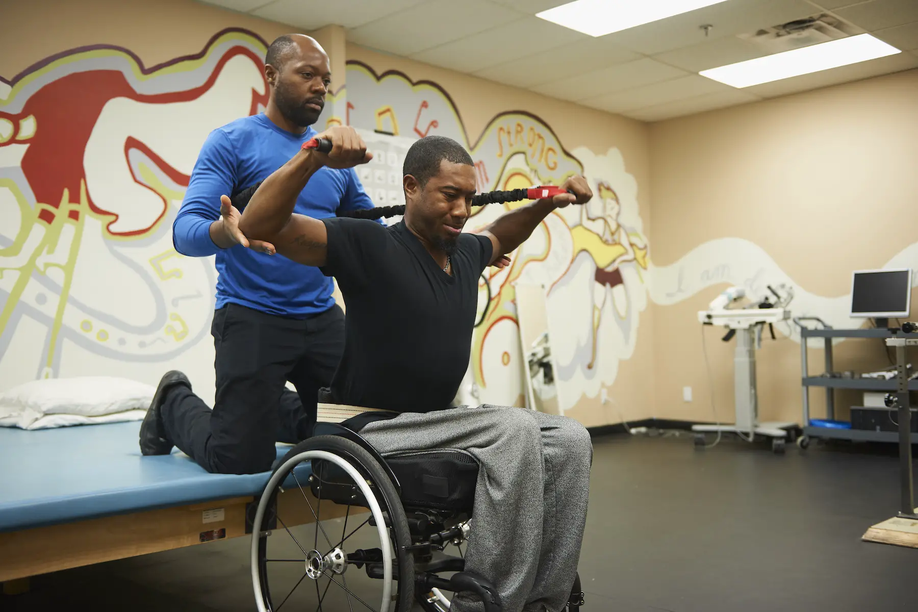 A physical therapist sits behind a man in a wheelchair, helping him strengthen his arms with a resistance band