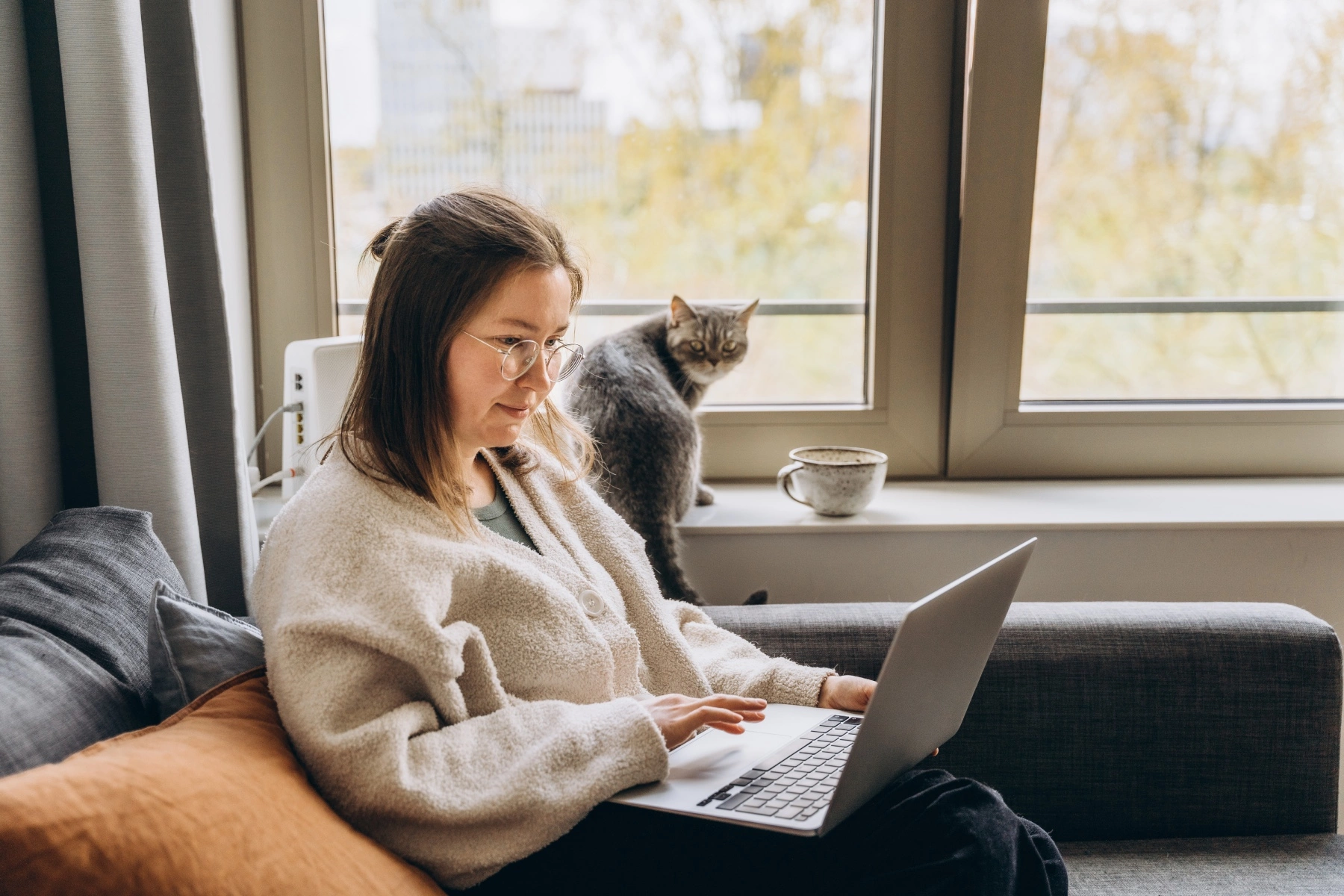 a woman sitting on a sofa at home working on her laptop as her cat sits on the window ledge behind her