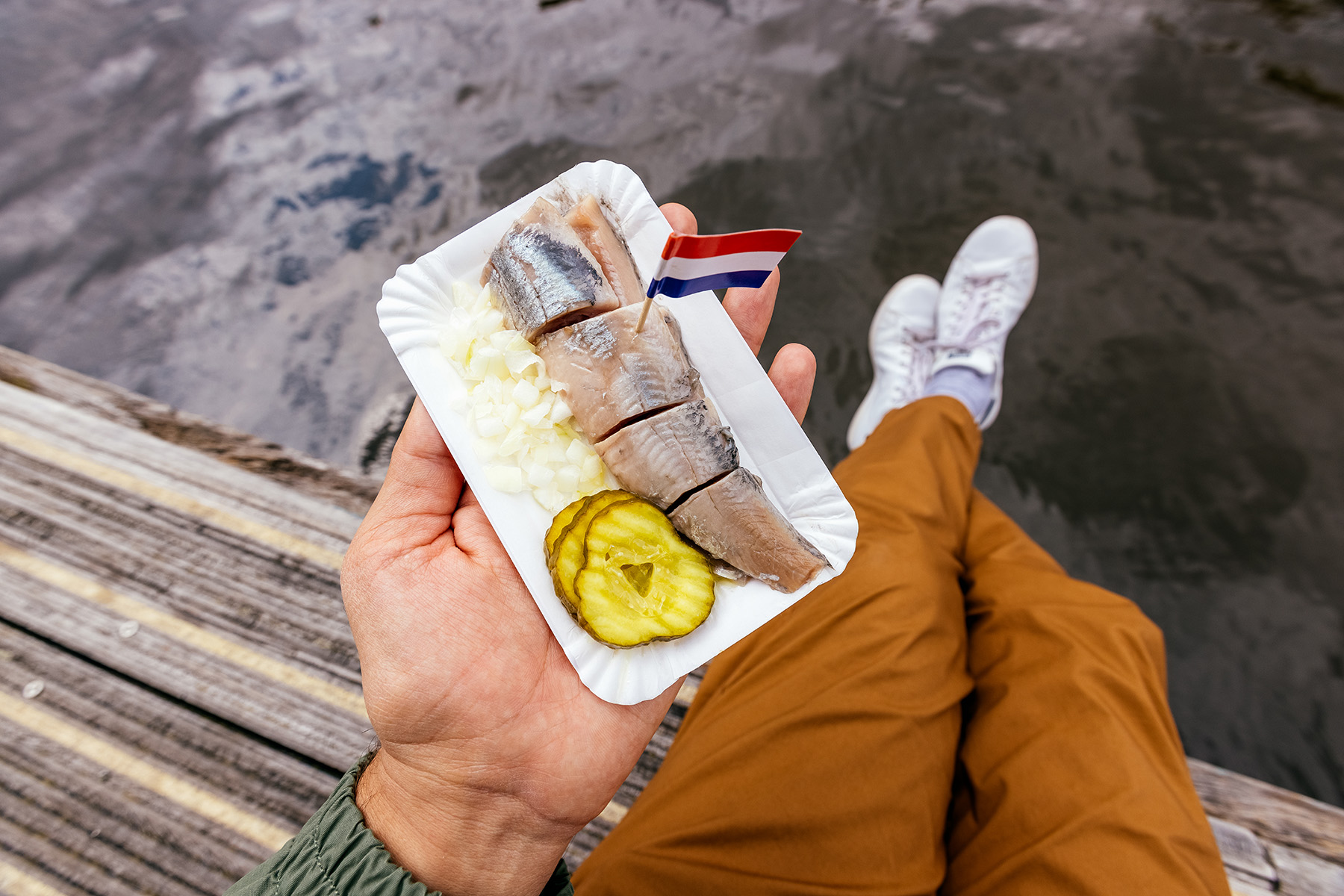 Person sitting by the side of the gracht, holding a paper plate with pieces of raw herring, onions, pickles, and a little Dutch flag.