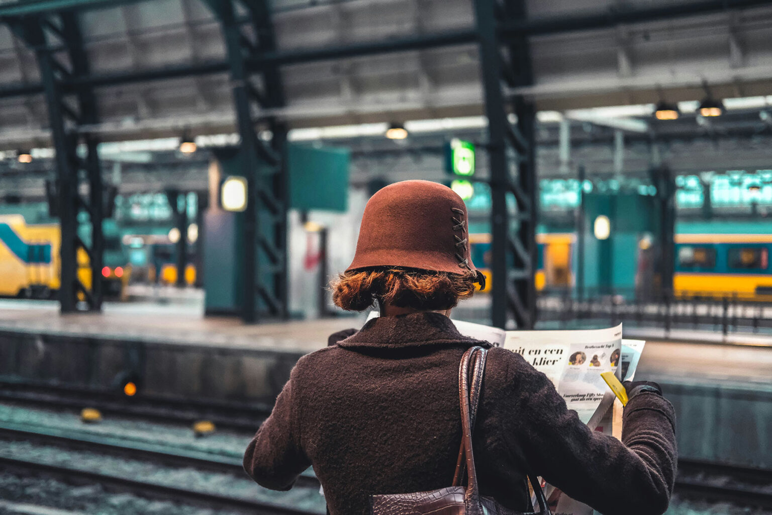 Woman with her back turned to the camera, reading a newspaper, while waiting on her train.