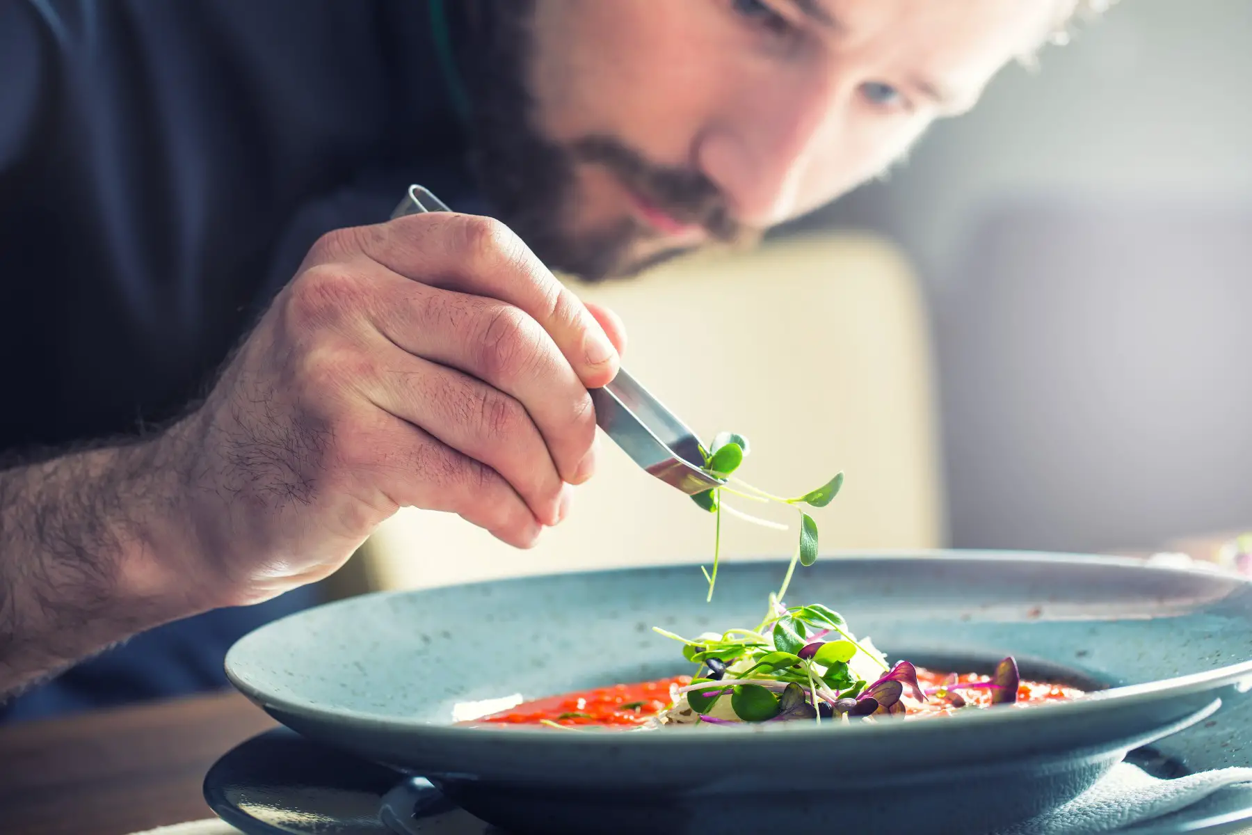 Chef in a restaurant putting herbs with some tweezers on tomato soup.