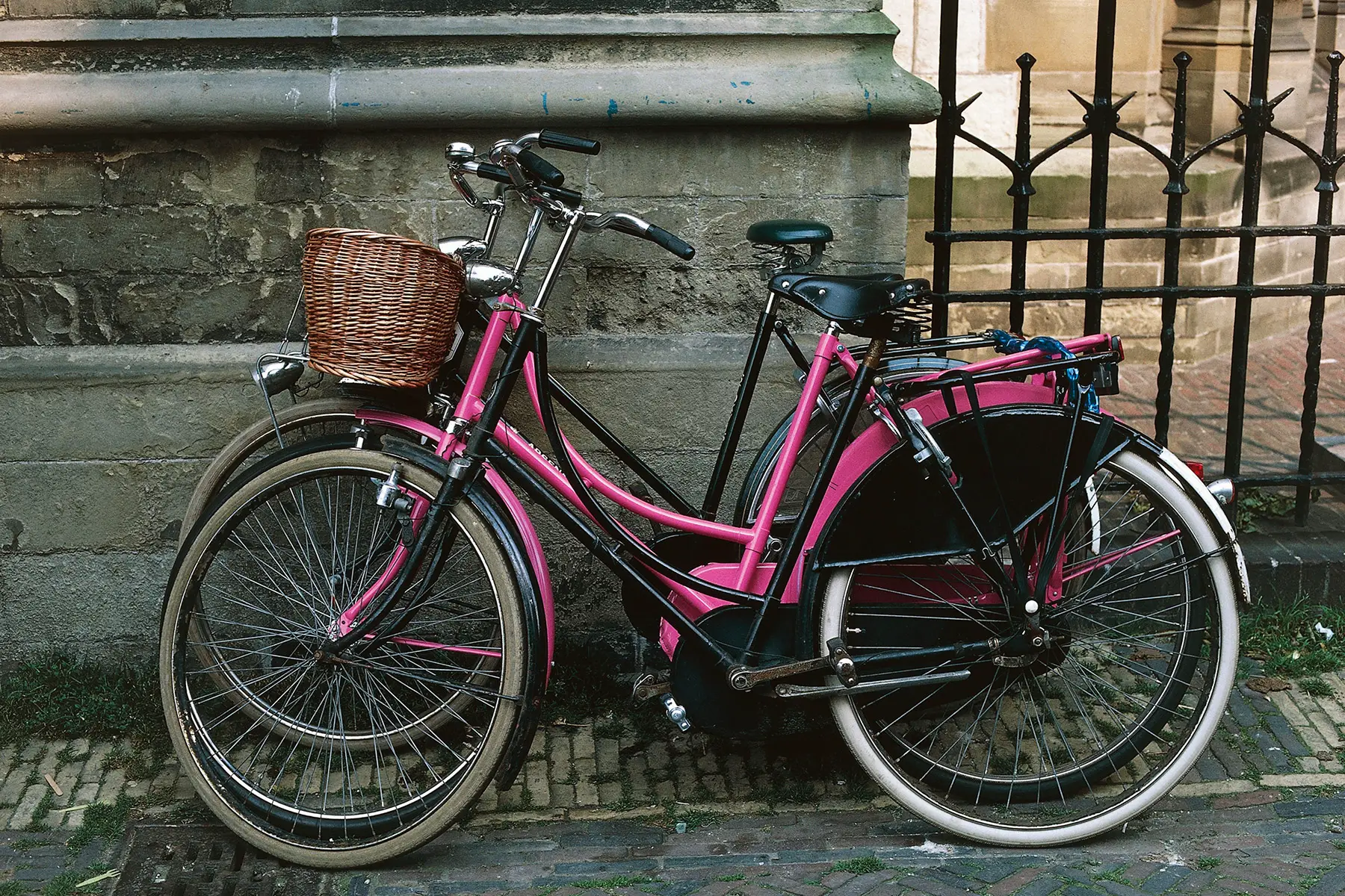 Two black bicycles and a pink one leaning against a wall.
