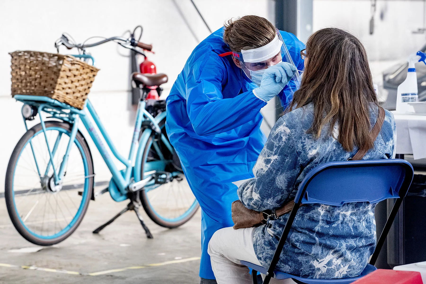 Medical professional with face mask using a COVID swap on a person sitting in front of them. In the background is a blue bike with a basket.
