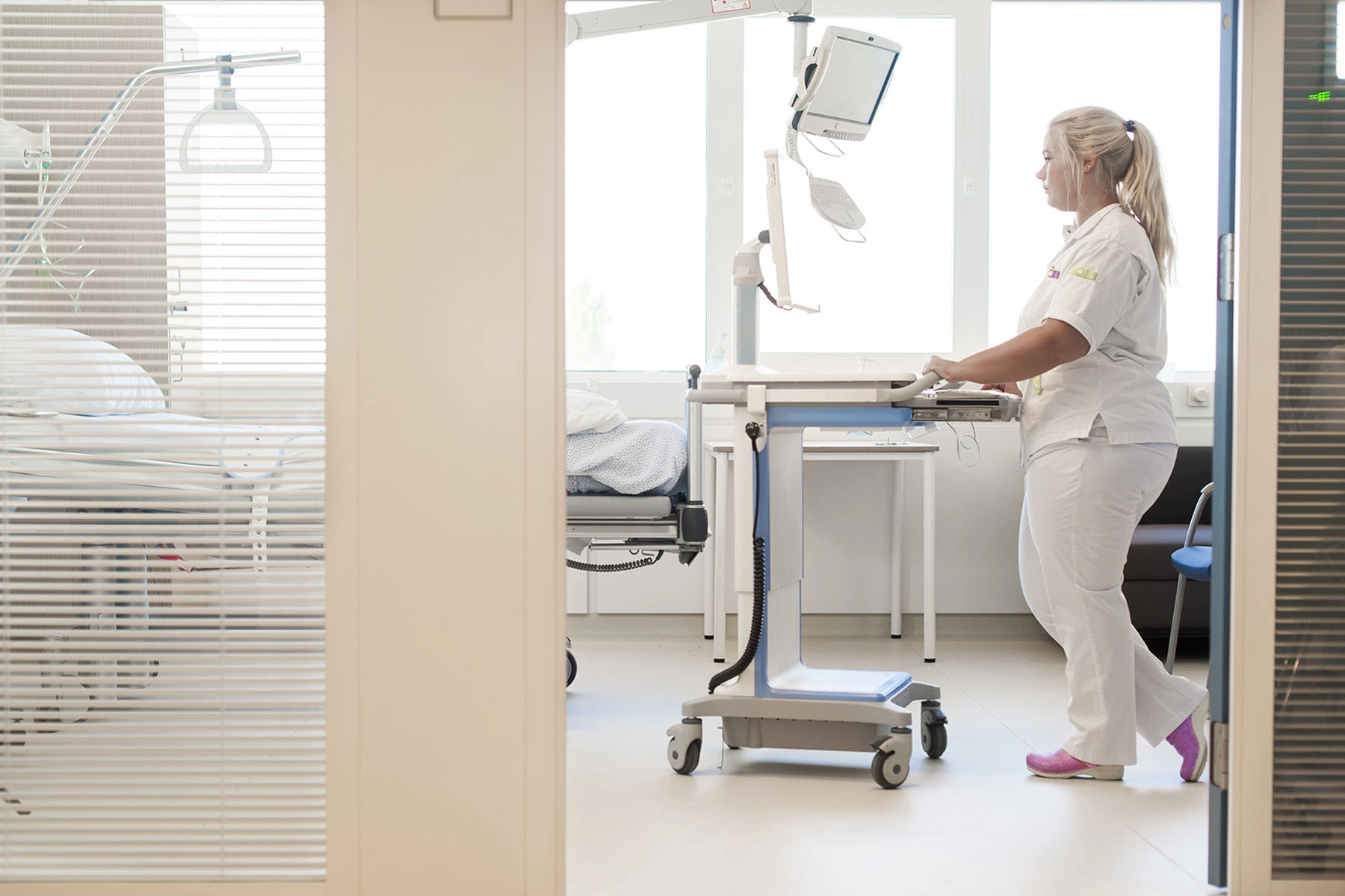 Nurse is wheeling a cart with a computer to an empty bedside.