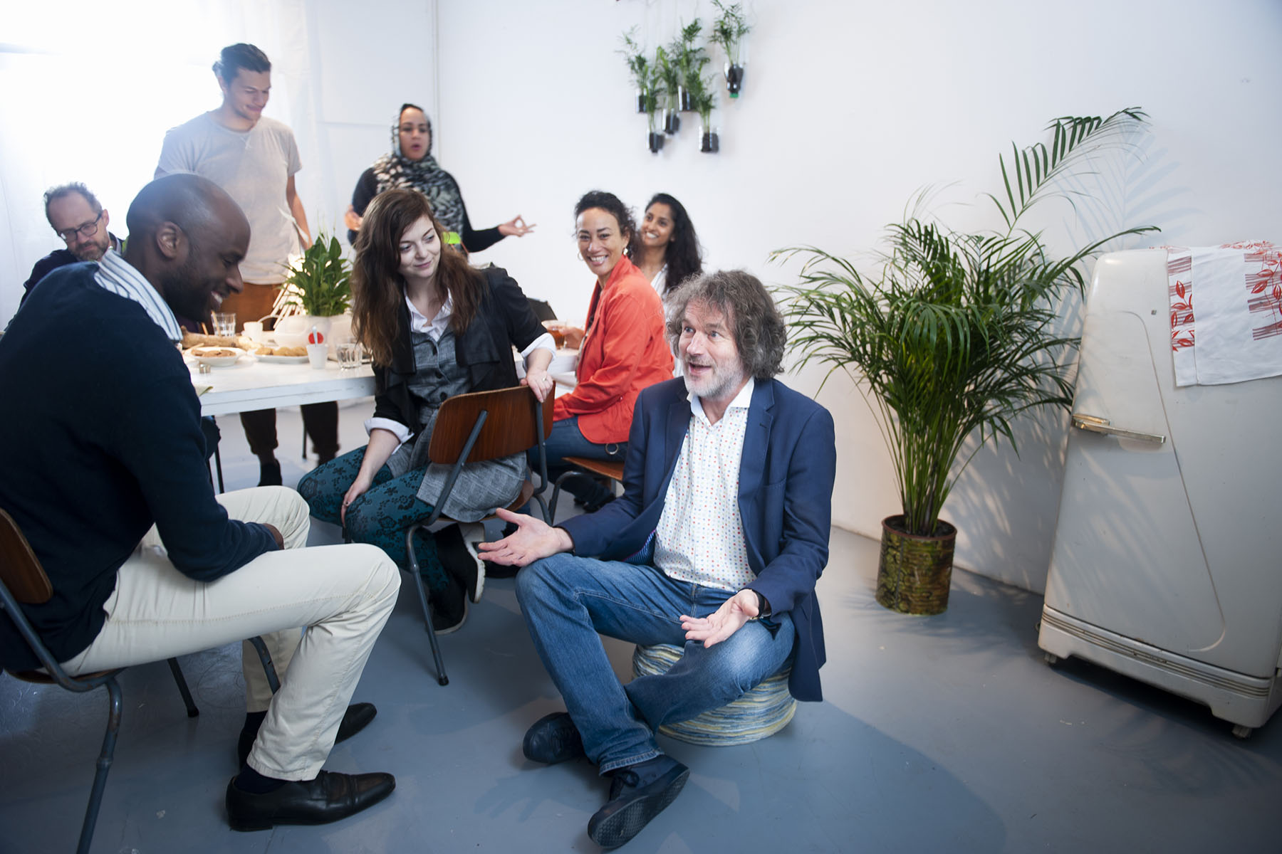 Group of colleagues of different levels sitting and standing around a table having a business meeting.  The CEO is sitting on a stool, being silly, and making the others laugh.