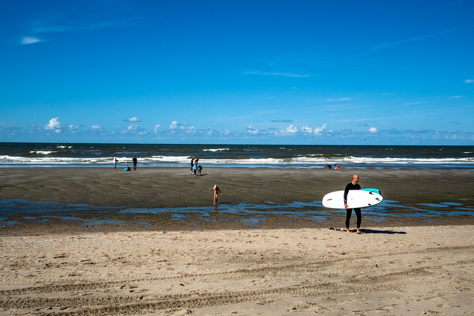 People are enjoying a sunny day at the beach of Hoek van Holland. One person is carrying a surf board.