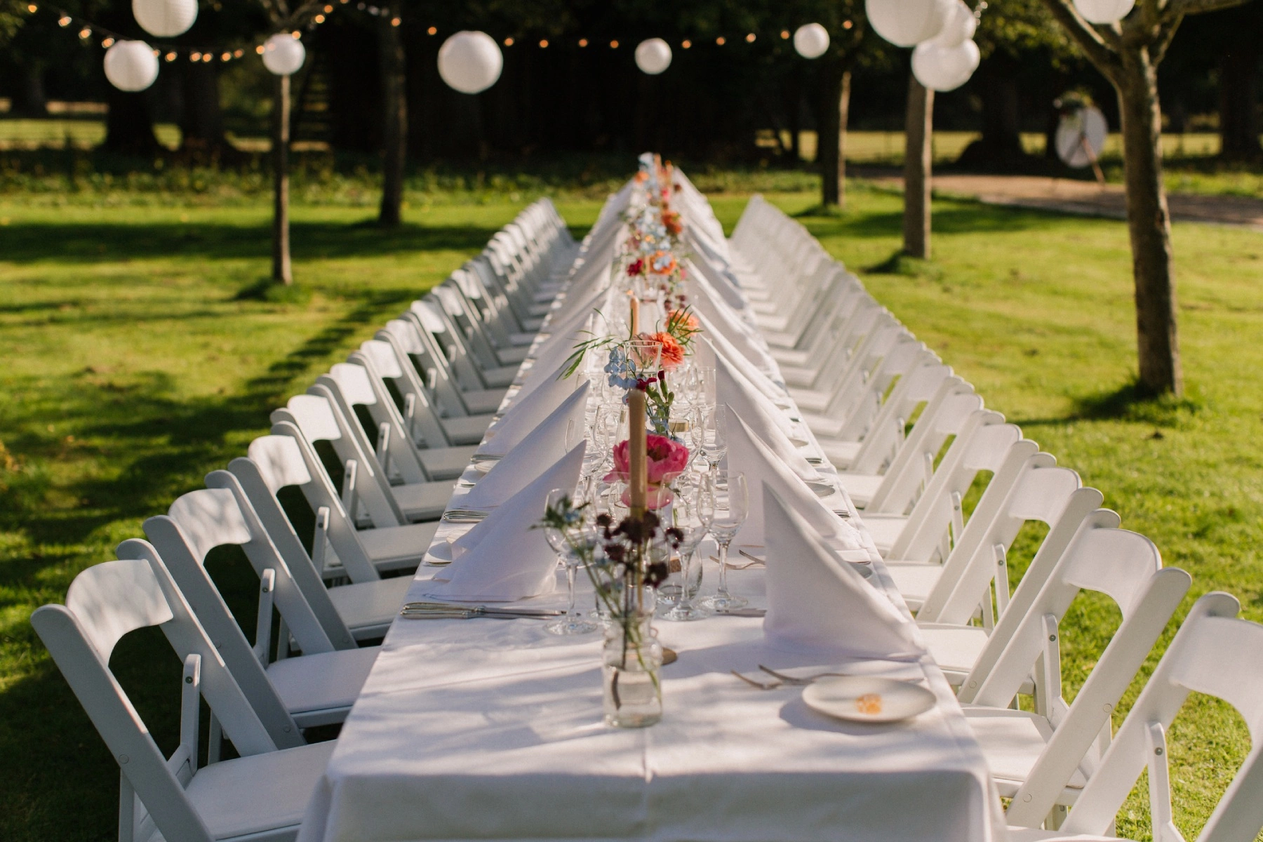 a beautifully styled wedding table decorated with pink flowers and white napkins sitting in the middle of a green lawn on a sunny afternoon
