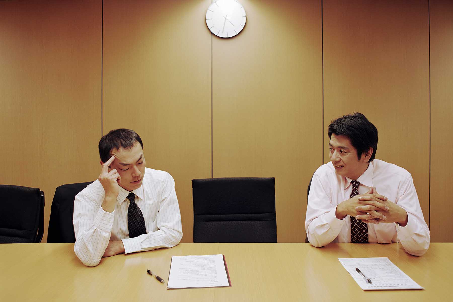 Two men sitting in an office with some very bad light. One is smiling looking at the other person, the other is pensive and looking down at the contract in front of him. 