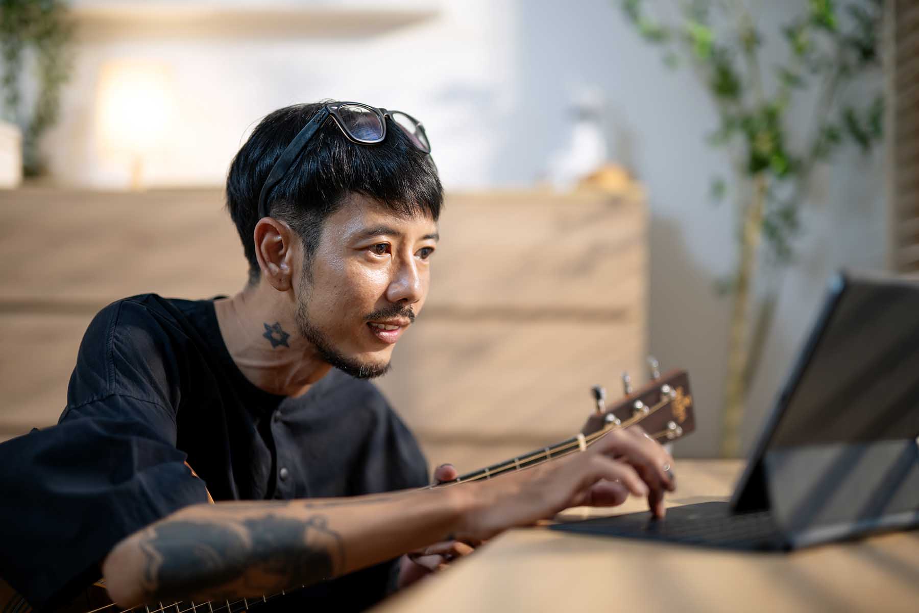 Smiling composer is sitting on the floor, holding his guitar on his lap, and writing down a music score on his laptop.