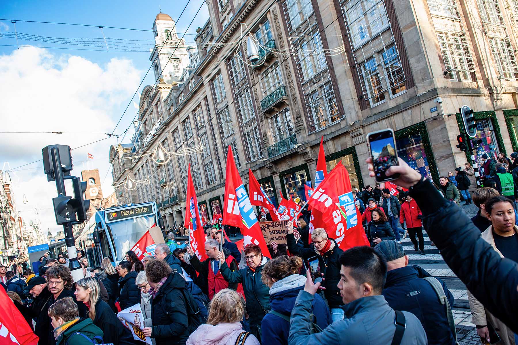 Protesters march in front of the department store De Bijenkorf in Amsterdam, the Netherlands, during a 2022 workers' strike. Employees took to the streets demanding higher wages and better conditions. Several workers carry red flags from the Netherlands Trade Union Confederation (FNV), which announced the strike.