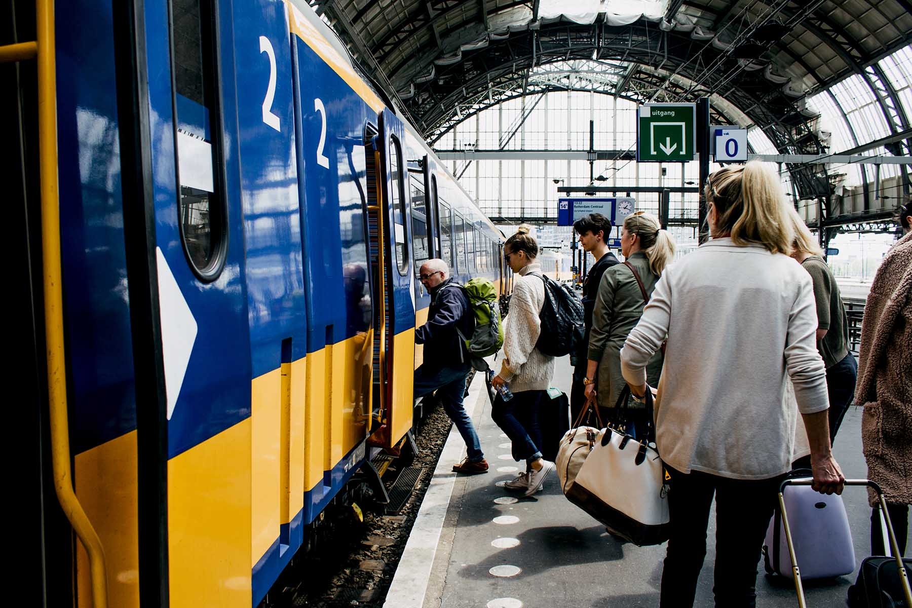 A group of people boarding the yellow NS train on a platform in Amsterdam, the Netherlands. Everyone is carrying large bags or traveling trollies.